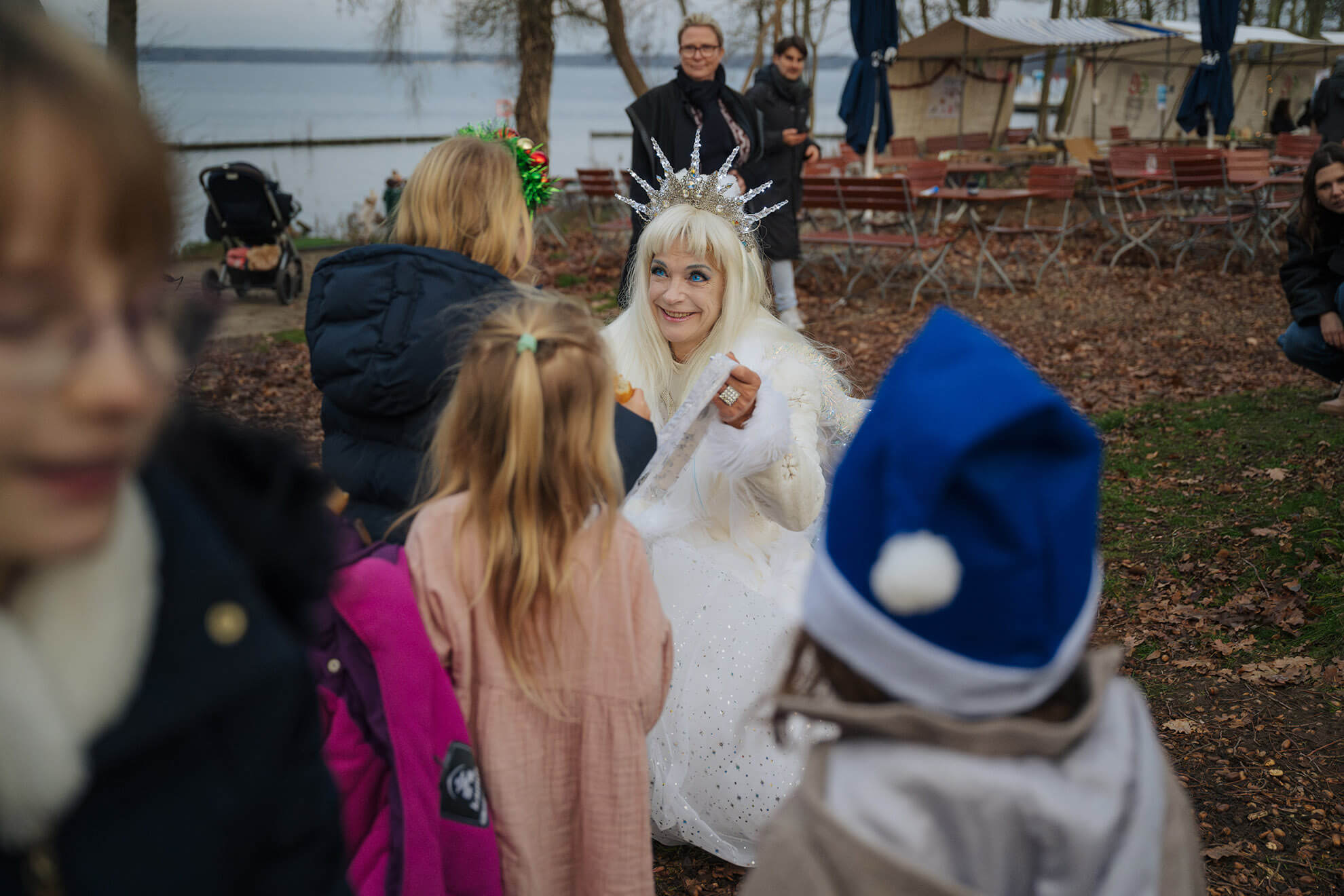 Cosmopolitanschool Berlin Eine Frau in einem weißen Kostüm mit Krone unterhält sich mit Kindern im Freien an einem See; im Hintergrund sind leere Stühle und Bäume zu sehen, und einige Kinder tragen festliche Hüte. loading=