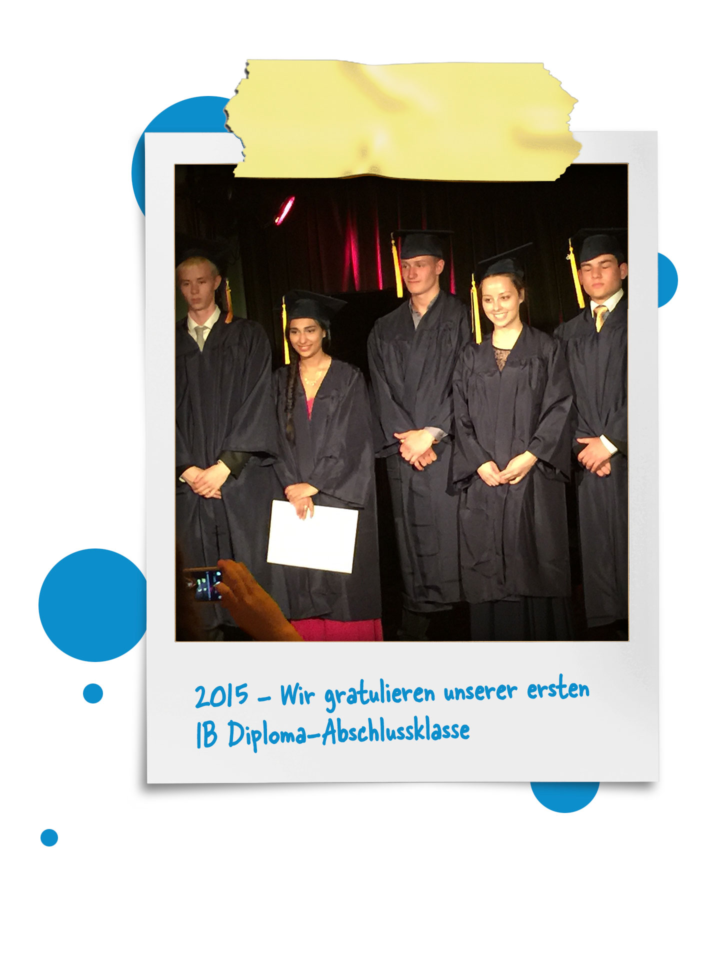 Cosmopolitanschool Berlin Fünf Schülerinnen und Schüler in schwarzen Abschlusskleidern und Mützen stehen auf der Bühne und halten ihre Diplome. Der Text darunter gratuliert dem ersten IB-Diplom-Abschlussjahrgang 2015 auf Deutsch. Blaue Punkte zieren den Rand des Fotos. loading=