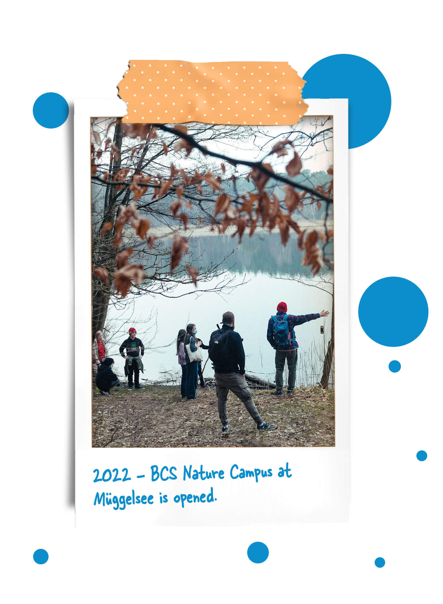 Cosmopolitanschool Berlin Eine Gruppe von Menschen steht in der Nähe des Ufers eines Sees, umgeben von Bäumen mit Herbstlaub, während eine Person auf das Wasser zeigt. Der Text lautet: 2022 - BCS Naturcampus am Müggelsee wird eröffnet. loading=