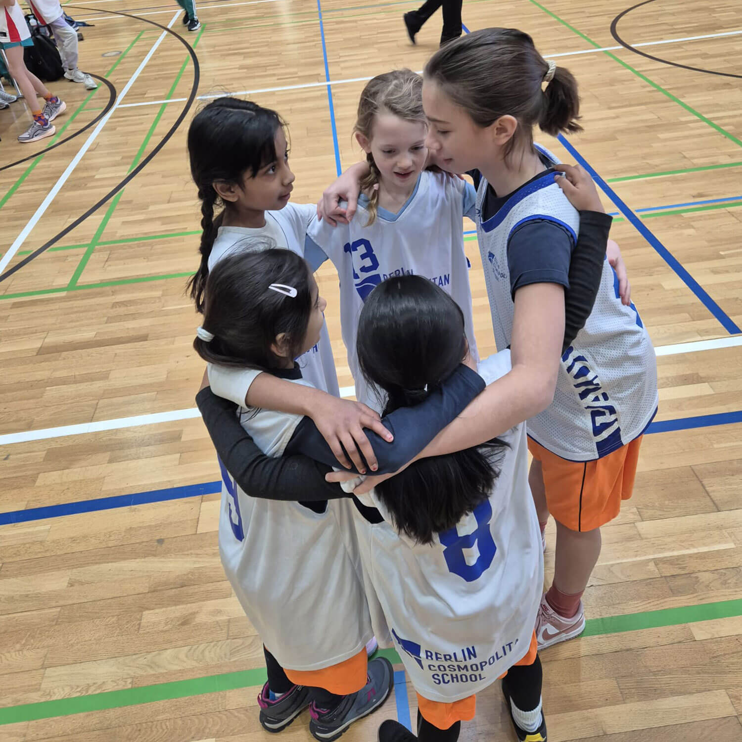 Cosmopolitanschool Berlin Fünf junge Mädchen in BCS-Grundschulmädchen-Basketballtrikots stehen vor ihrem Spiel in der ALBA-Grundschulliga 2026 auf dem Hallenplatz, legen die Arme umeinander, lächeln und reden. Der Platz hat einen Holzboden mit grünen und weißen Linien. loading=