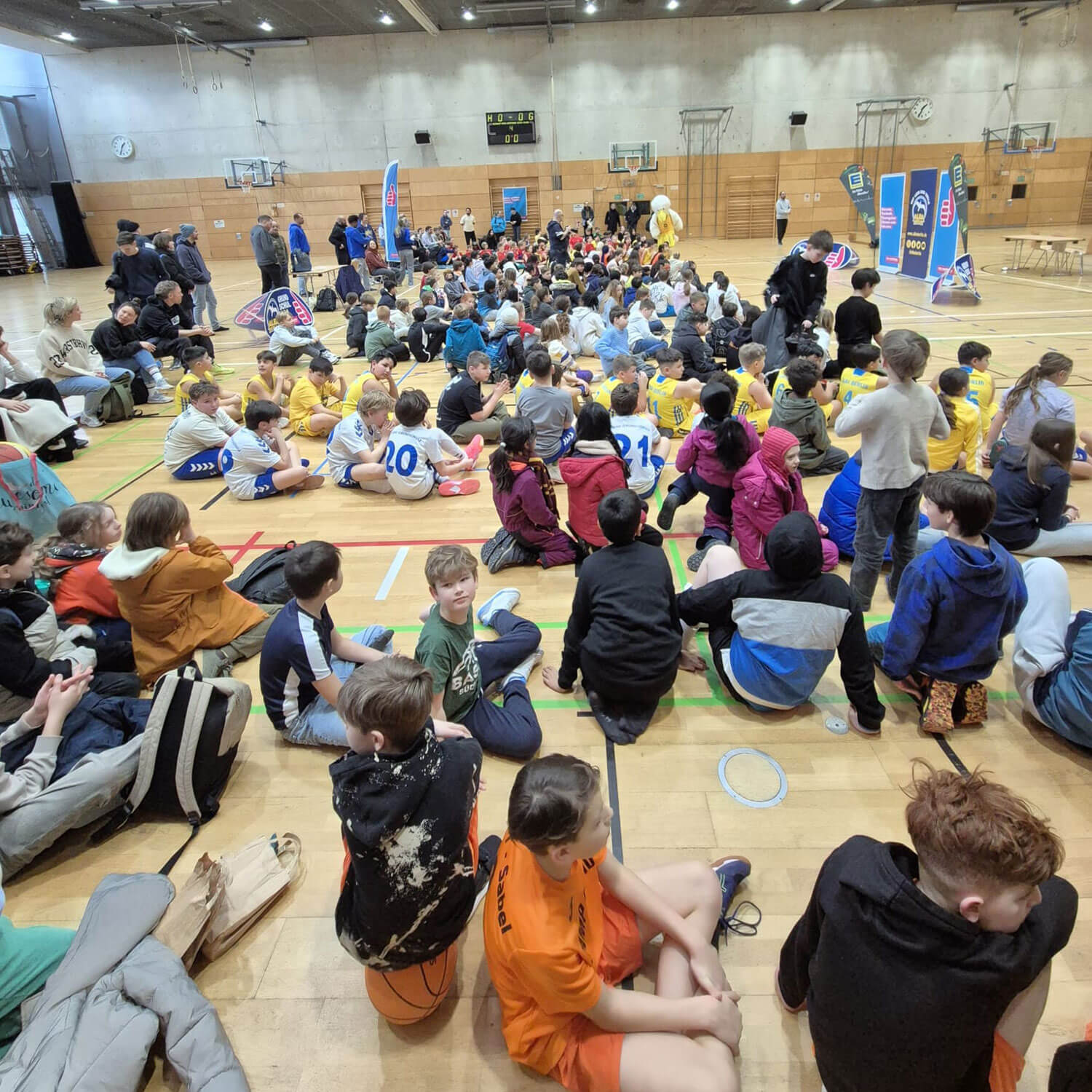 Cosmopolitanschool Berlin Eine große Gruppe von Kindern und Erwachsenen sitzt auf dem Boden der Sporthalle, gegenüber einer Bühne. Viele tragen Sportuniformen der BCS-Grundschulmädchen 2026 ALBA-Grundschulliga, und einige halten Taschen oder Bälle. Auf dem Holzboden liegen Sportgeräte. loading=