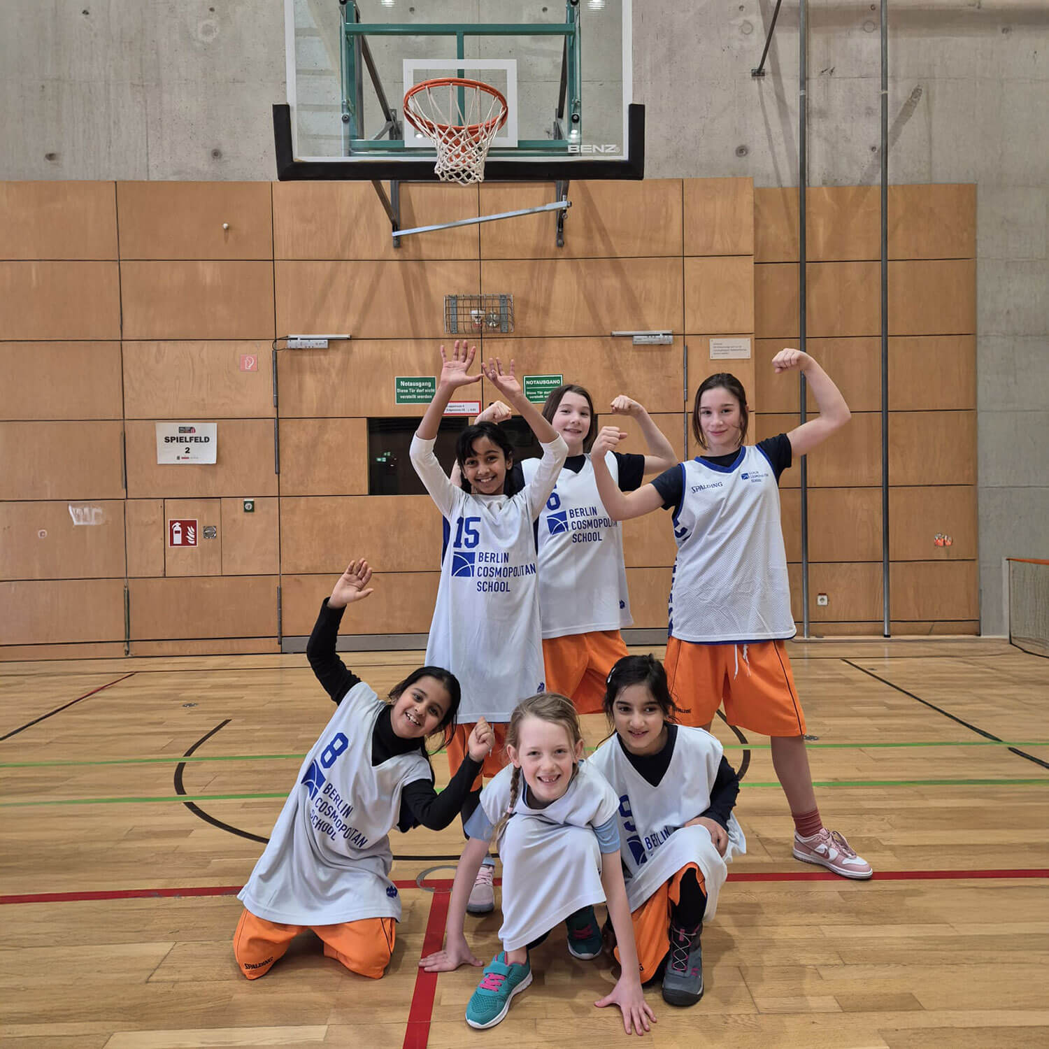 Cosmopolitanschool Berlin Sechs junge Mädchen in Basketballuniformen posieren fröhlich unter einem Basketballkorb in einer Turnhalle und vertreten die BCS-Grundschulmädchen in der ALBA-Grundschulliga 2026. Einige strecken ihre Arme aus, andere knien nieder, alle lächeln und zeigen Teamgeist. loading=