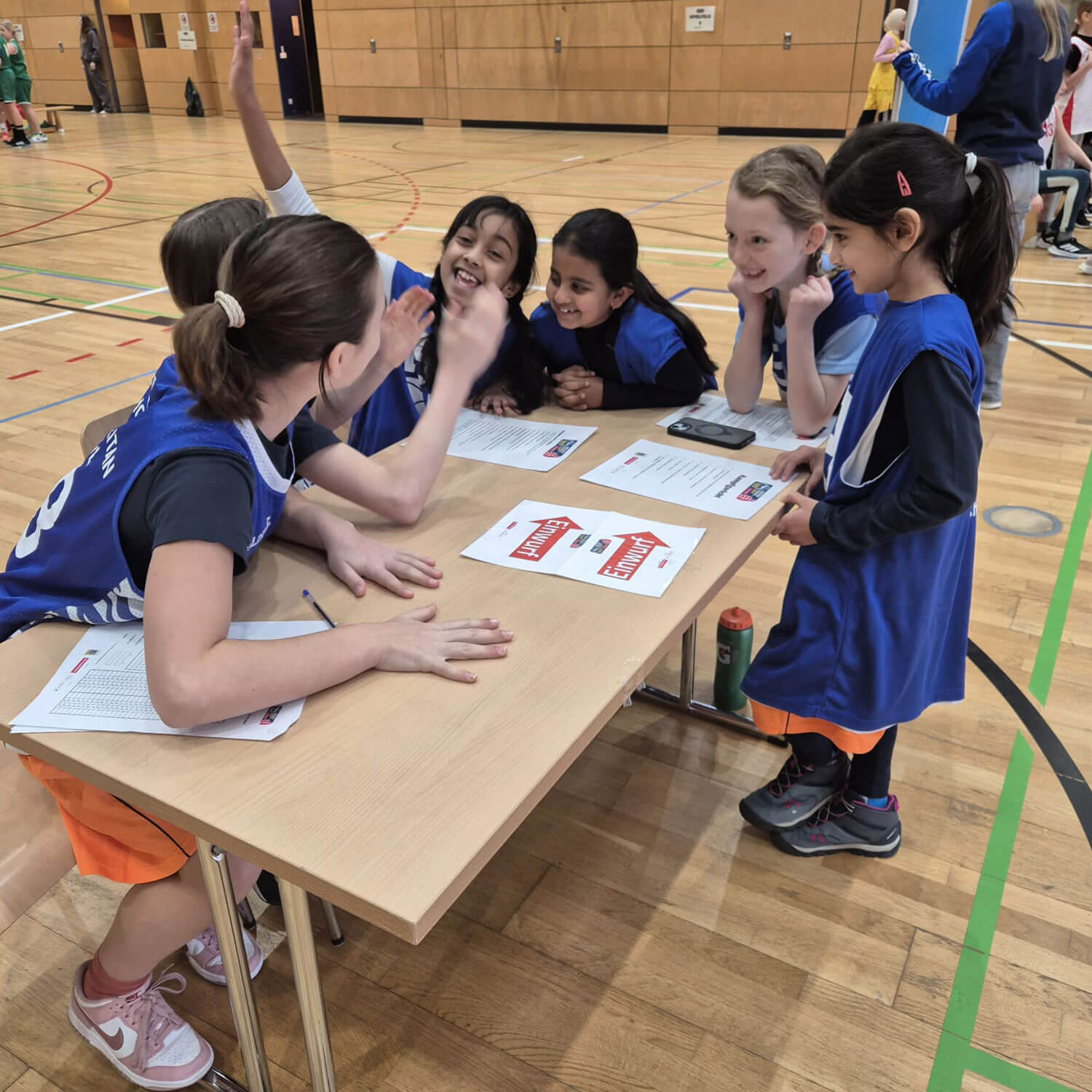 Cosmopolitanschool Berlin Sechs junge Mädchen in blauen Sportuniformen aus der ALBA-Grundschulliga sitzen und stehen in einer Turnhalle um einen Tisch herum, lächeln und geben sich die Hand, während sie auf Papiere und ein Telefon schauen, und fangen so den Geist der Teamarbeit und die Begeisterung für den Mädchensport ein.