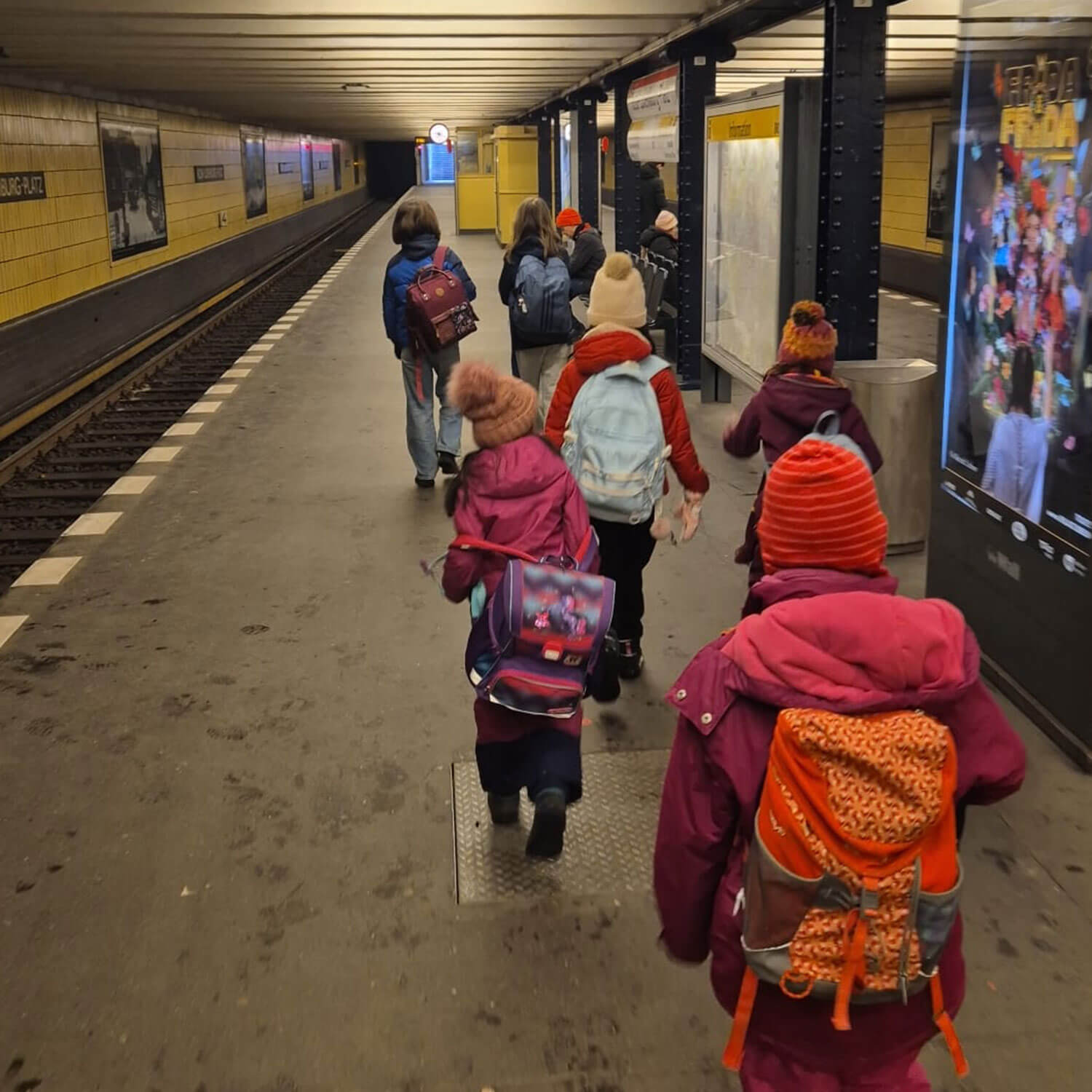Cosmopolitanschool Berlin Eine Gruppe junger Kinder, darunter auch Mädchen der BCS-Grundschule, gehen in bunter Winterkleidung und mit Rucksäcken an einem U-Bahn-Bahnsteig entlang. Auf der rechten Seite sind die gelb gekachelten Bahnhofswände und die Plakate der ALBA-Grundschulliga zu sehen.