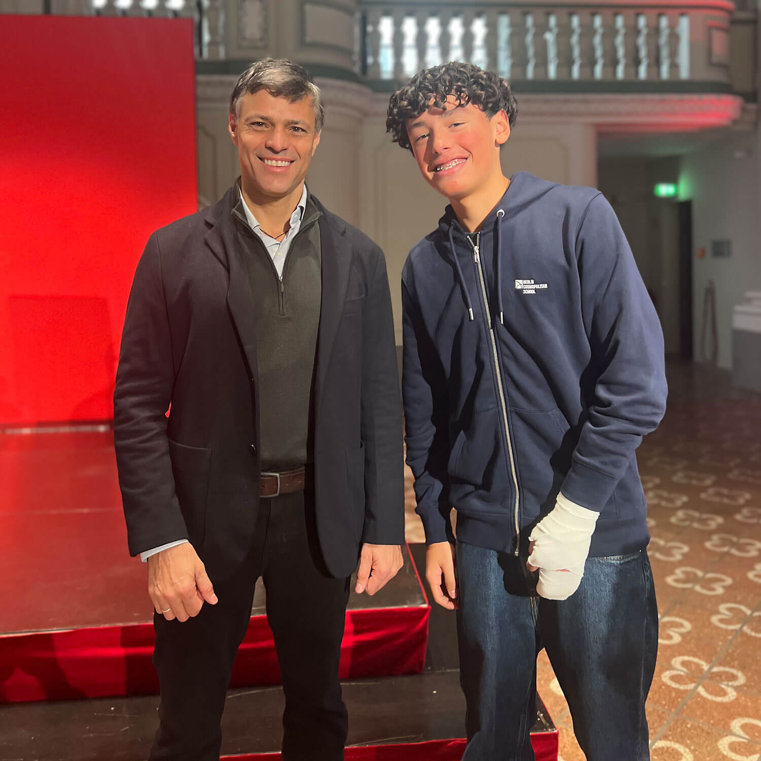 Cosmopolitanschool Berlin Zwei Personen stehen in einem Innenraum auf einem gemusterten Boden vor einer roten Wand. Beide lächeln - der eine in einer schwarzen Jacke, der andere in einem marineblauen Kapuzenpulli und Jeans mit einer bandagierten Hand - als würden sie für einen automatisch gespeicherten Entwurf posieren.