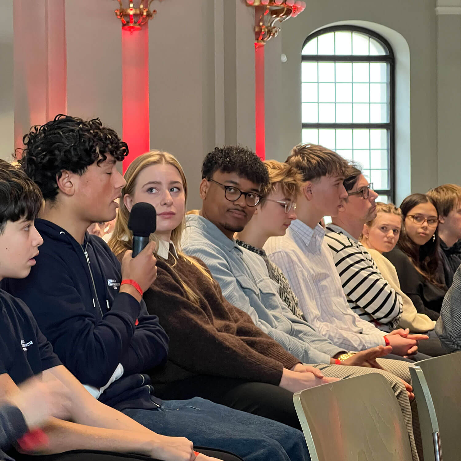 Cosmopolitanschool Berlin Eine Gruppe junger Menschen sitzt in Reihen in einem Raum mit hohen Fenstern und rot beleuchteten Säulen. Eine Person in der ersten Reihe hält ein Mikrofon und spricht, während die anderen, die vielleicht einen automatisch gespeicherten Entwurf durchgehen, aufmerksam zuhören.
