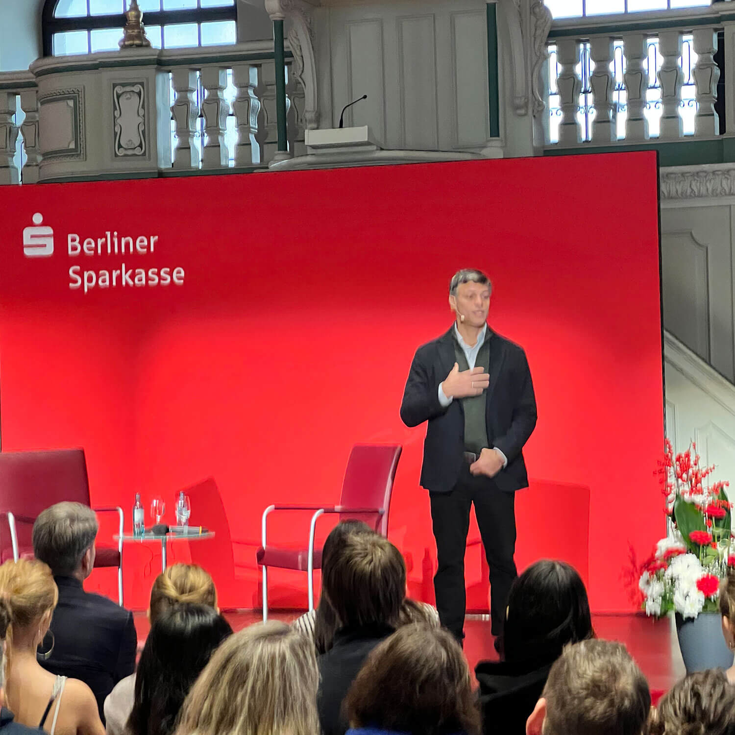 Cosmopolitanschool Berlin Ein Mann steht auf der Bühne vor einem roten Hintergrund mit dem Logo der Berliner Sparkasse und spricht zu einem Publikum. Auf der Bühne stehen zwei leere Stühle und ein Tisch mit Wasserflaschen, rechts daneben Blumen.