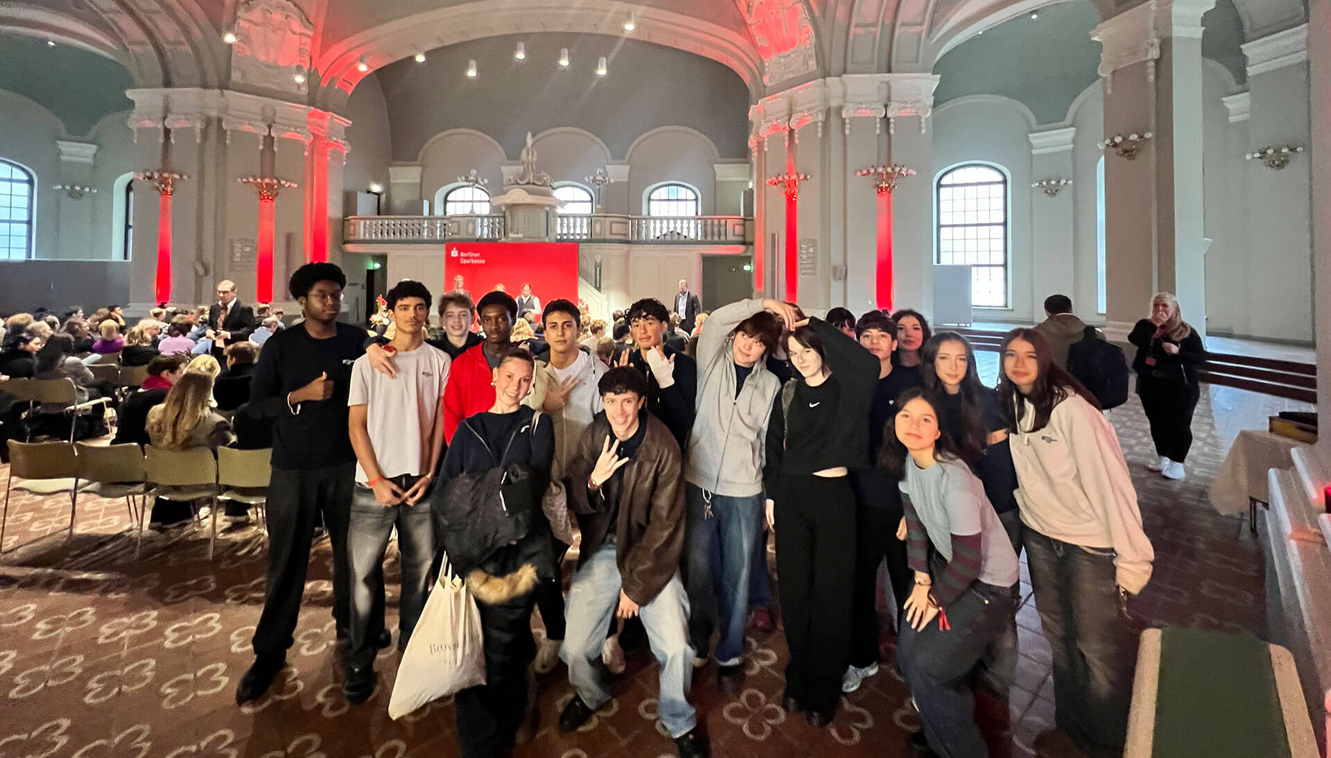 Cosmopolitanschool Berlin Eine Gruppe junger Leute posiert zusammen und lächelt in einem großen Saal mit hohen Decken, verzierten Bögen und roter Beleuchtung. Ein Publikum sitzt auf Stühlen vor einer Bühne im Hintergrund. loading=