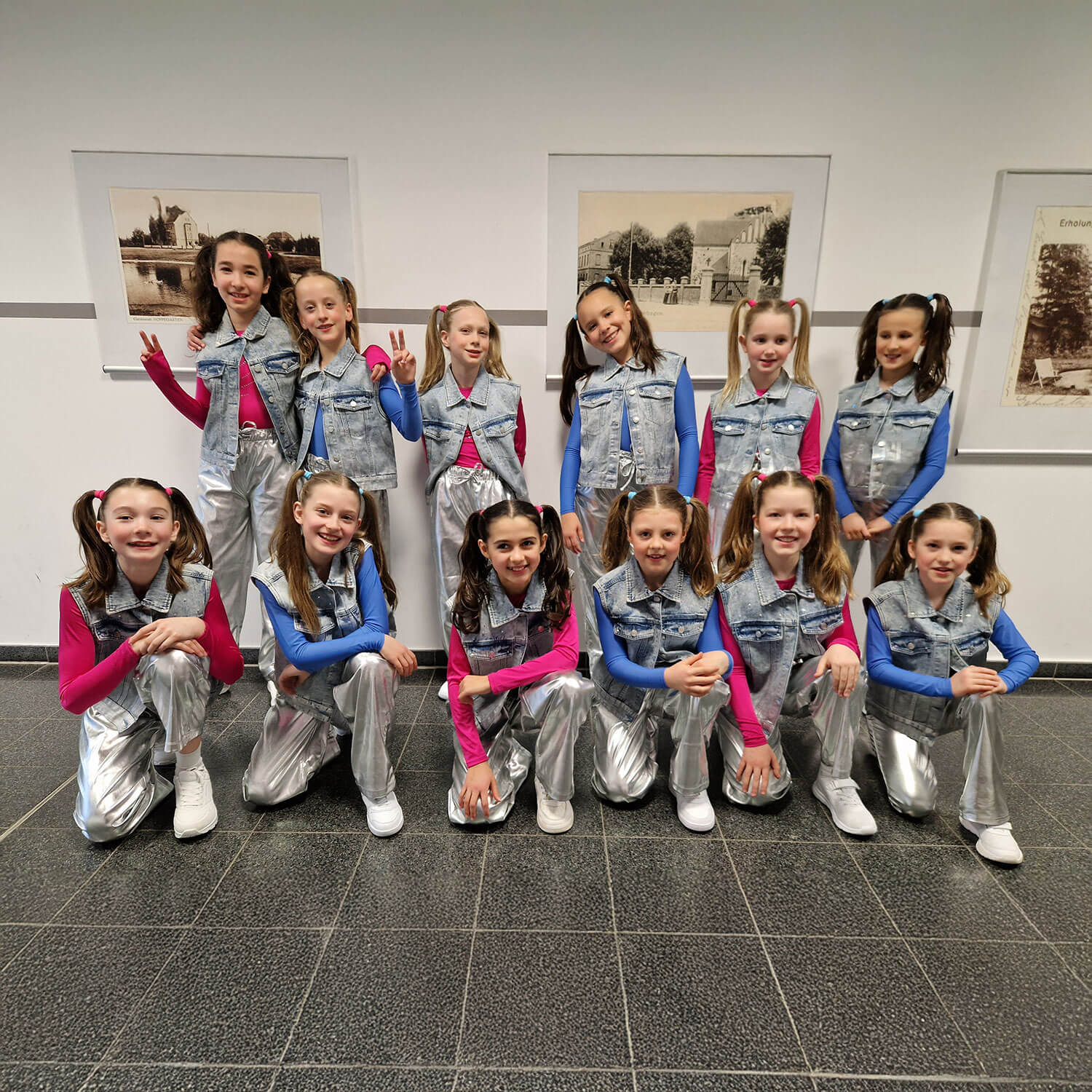 Cosmopolitanschool Berlin Zwölf junge Mädchen in passender Kleidung - silberne Hosen, blaue und rosafarbene Oberteile und Jeanswesten - posieren lächelnd zusammen in einem Raum. Die Gruppe steht und kniet in zwei Reihen vor einem gerahmten Wandbild. loading=