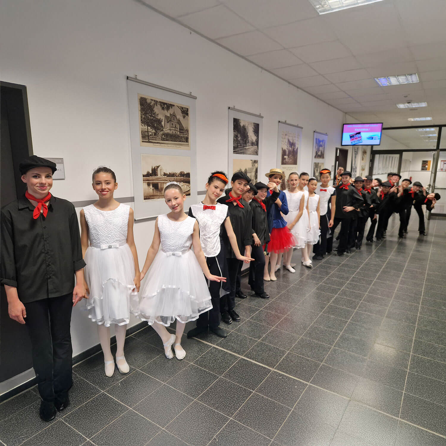 Cosmopolitanschool Berlin Eine Gruppe junger Tänzerinnen und Tänzer in Kostümen, einige in weißen Kleidern mit roten Akzenten, andere in schwarzen Kostümen mit roten Schals, stehen in einem mit gerahmten Bildern gesäumten Flur, halten sich an den Händen und lächeln in die Kamera.