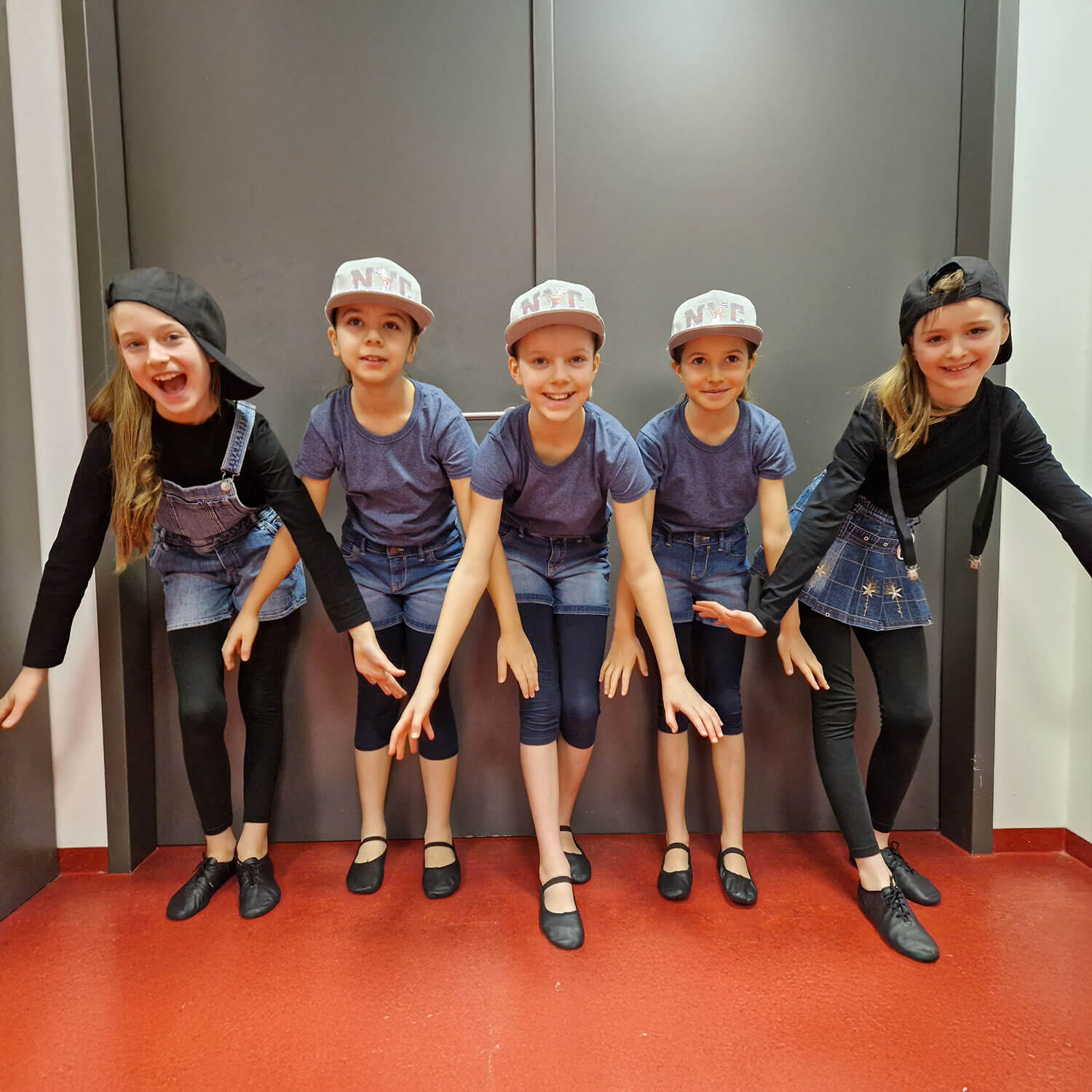 Cosmopolitanschool Berlin Fünf junge Mädchen in passender Tanzkleidung - blaue Oberteile, Jeansröcke, schwarze Leggings und schwarze Schuhe - posieren vor einer grauen Wand. Drei tragen helle Mützen, zwei dunkle. Alle lächeln, die Arme nach vorne gestreckt. loading=