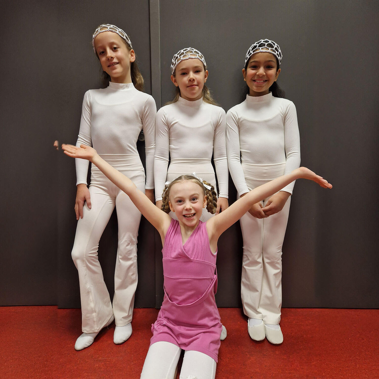 Cosmopolitanschool Berlin Vier Kinder posieren in einem Haus. Drei von ihnen stehen in passenden weißen Kleidern und Stirnbändern und lächeln nebeneinander. Ein Kind in einem rosafarbenen Outfit kniet mit ausgestreckten Armen davor und lächelt in die Kamera.