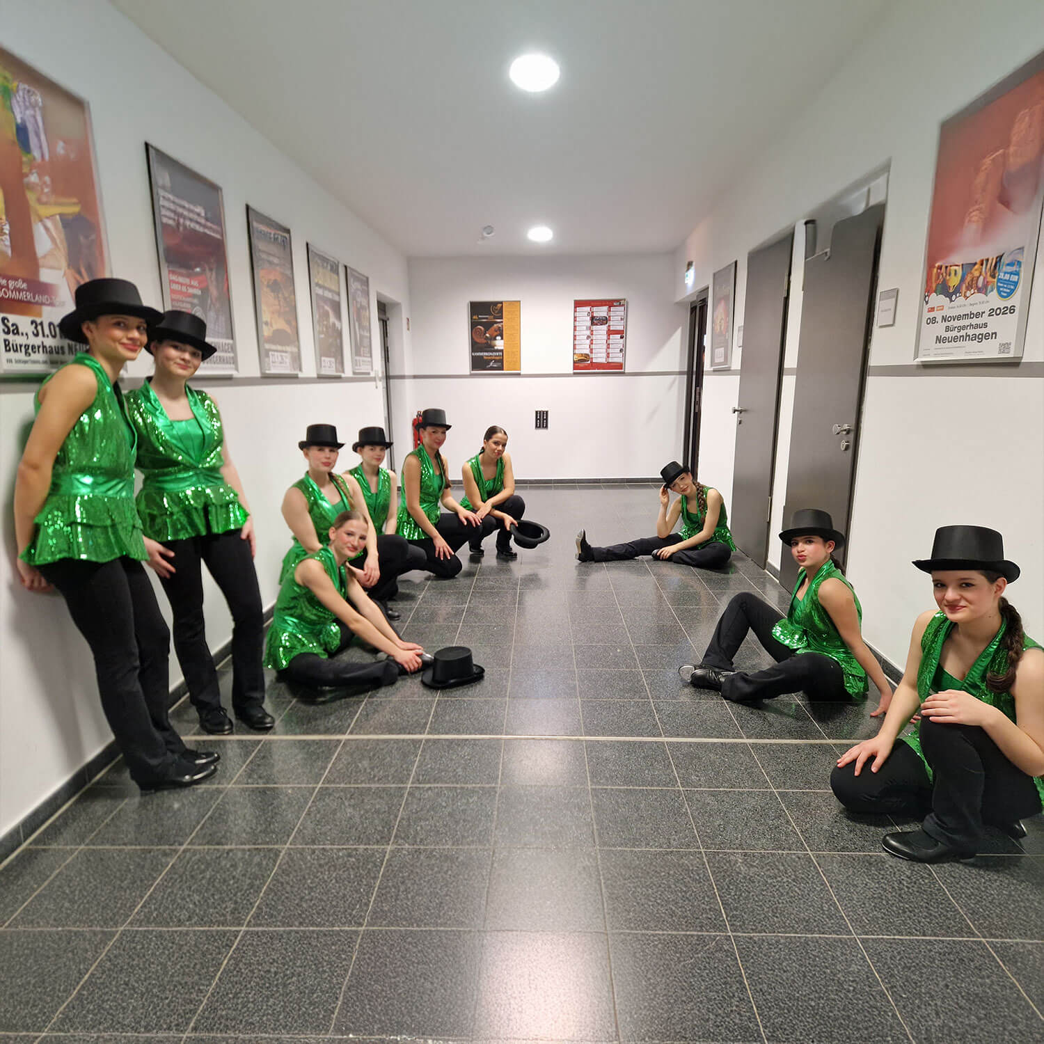 Cosmopolitanschool Berlin Eine Gruppe von Tänzern in grünen Paillettenoberteilen, schwarzen Hosen und schwarzen Hüten posiert in einem Flur. Einige stehen auf der linken Seite, während andere auf dem Boden entlang der beiden Wände sitzen und in die Kamera lächeln. An den Wänden befinden sich Werbeplakate. loading=