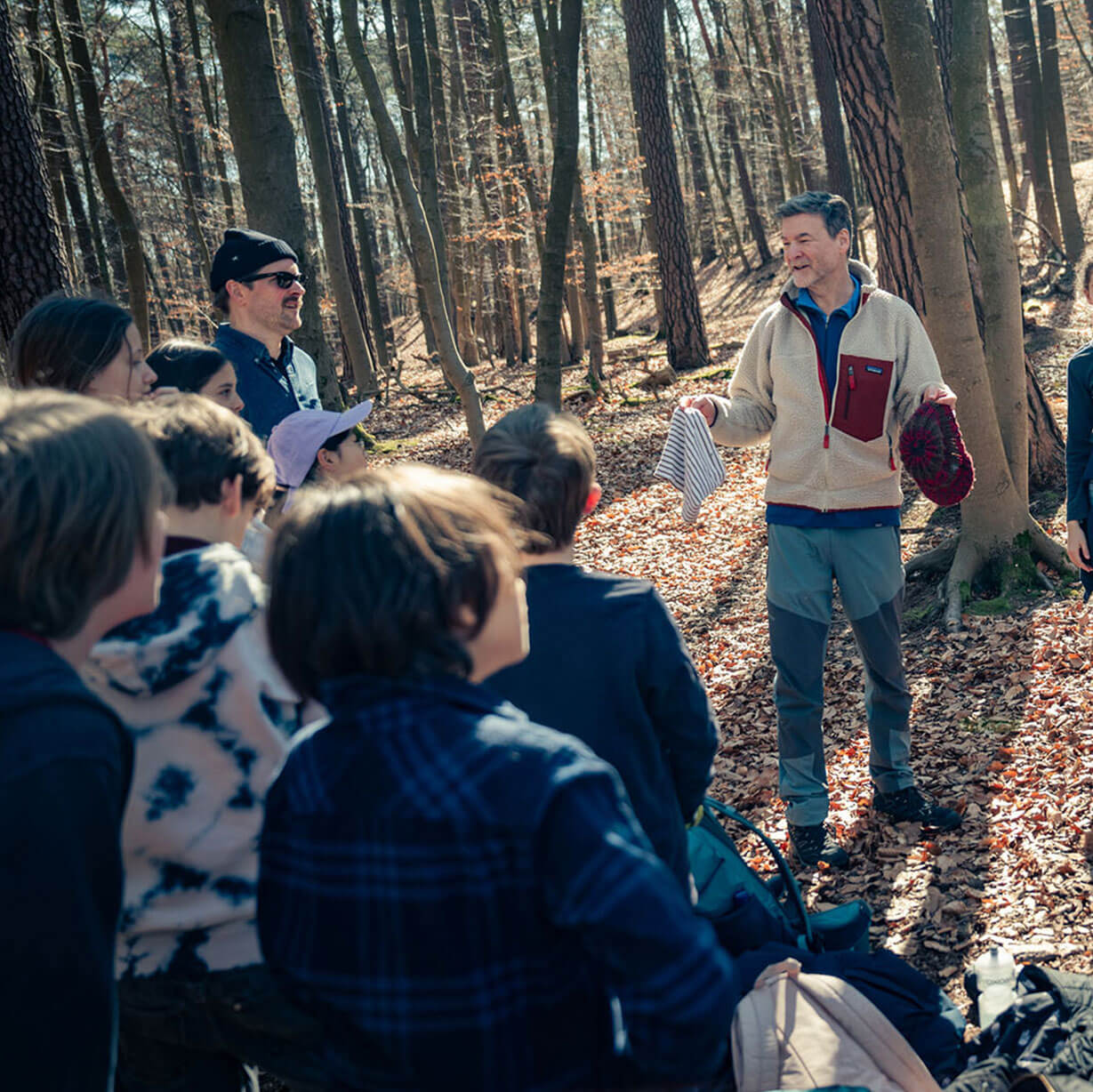 Cosmopolitanschool Berlin Eine Gruppe von Menschen, darunter Kinder und Erwachsene, ist in einem sonnenbeschienenen Wald versammelt und hört einem älteren Mann zu, der spricht und Gegenstände in seinen Händen hält. Der Boden ist mit abgefallenen Blättern bedeckt. loading=