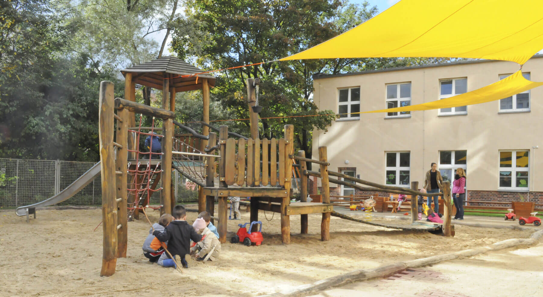 Cosmopolitanschool Berlin Kinder spielen auf einem Holzspielplatz mit Rutschen, Seilen und Sand unter großen gelben Sonnensegeln im Kindergarten und in der Vorschule. Mehrere Erwachsene beaufsichtigen sie in der Nähe, und im Hintergrund ist ein beiges Gebäude zu sehen.