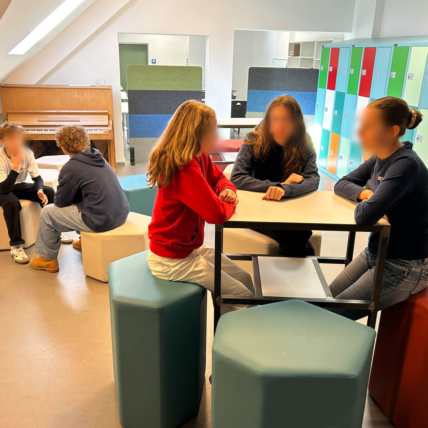 Cosmopolitanschool Berlin Mehrere Schüler sitzen in Gruppen in einem hell erleuchteten Klassenzimmer mit bunten Spinden, modernen Möbeln und einem Klavier im Hintergrund und unterhalten sich. Die Gesichter sind zum Schutz der Privatsphäre unscharf.