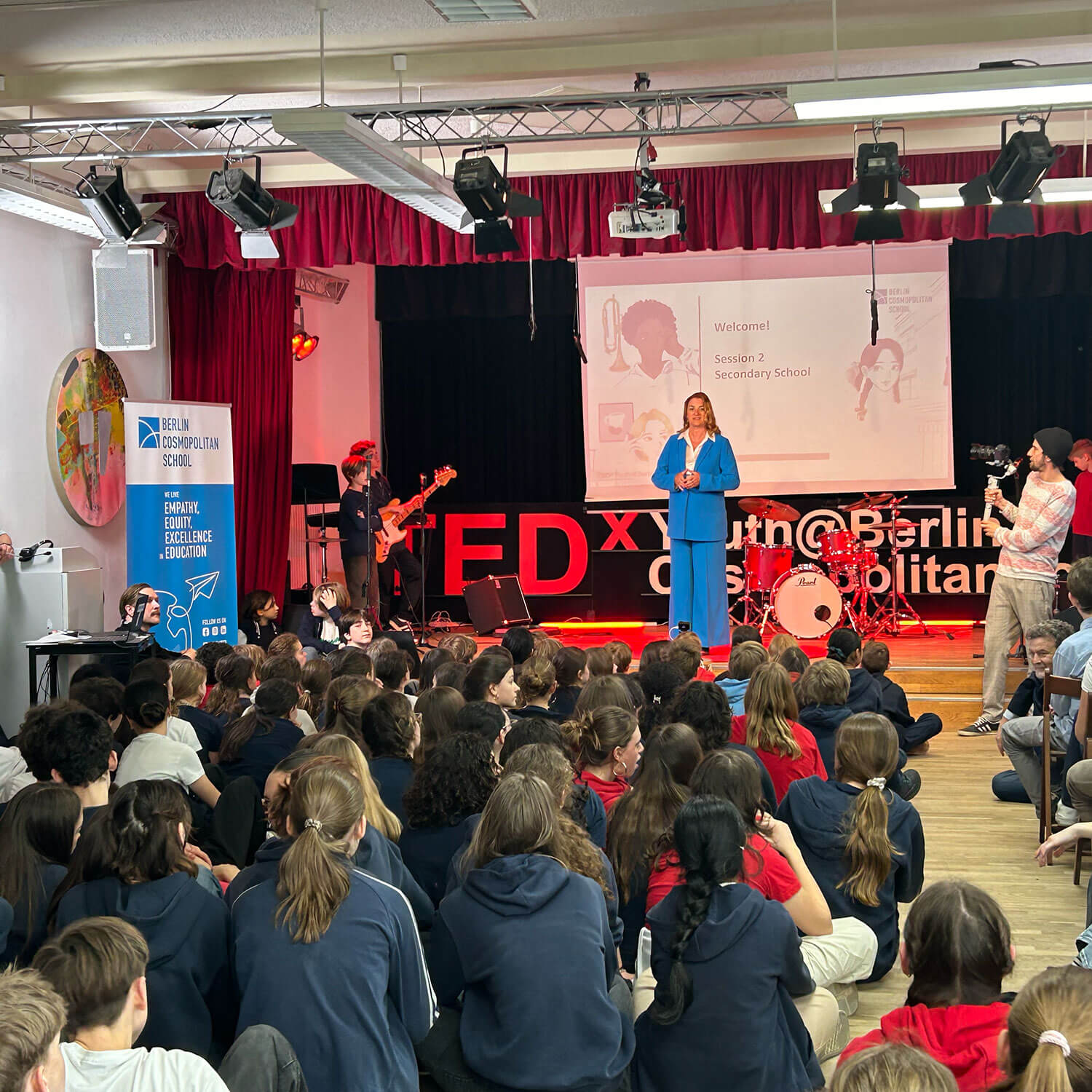 Cosmopolitanschool Berlin Eine Frau in einem blauen Anzug spricht auf der Bühne einer TEDx-Veranstaltung in einer Schulaula, während links eine Band spielt und ein Kameramann filmt. Schüler sitzen auf dem Boden und schauen aufmerksam zu.
