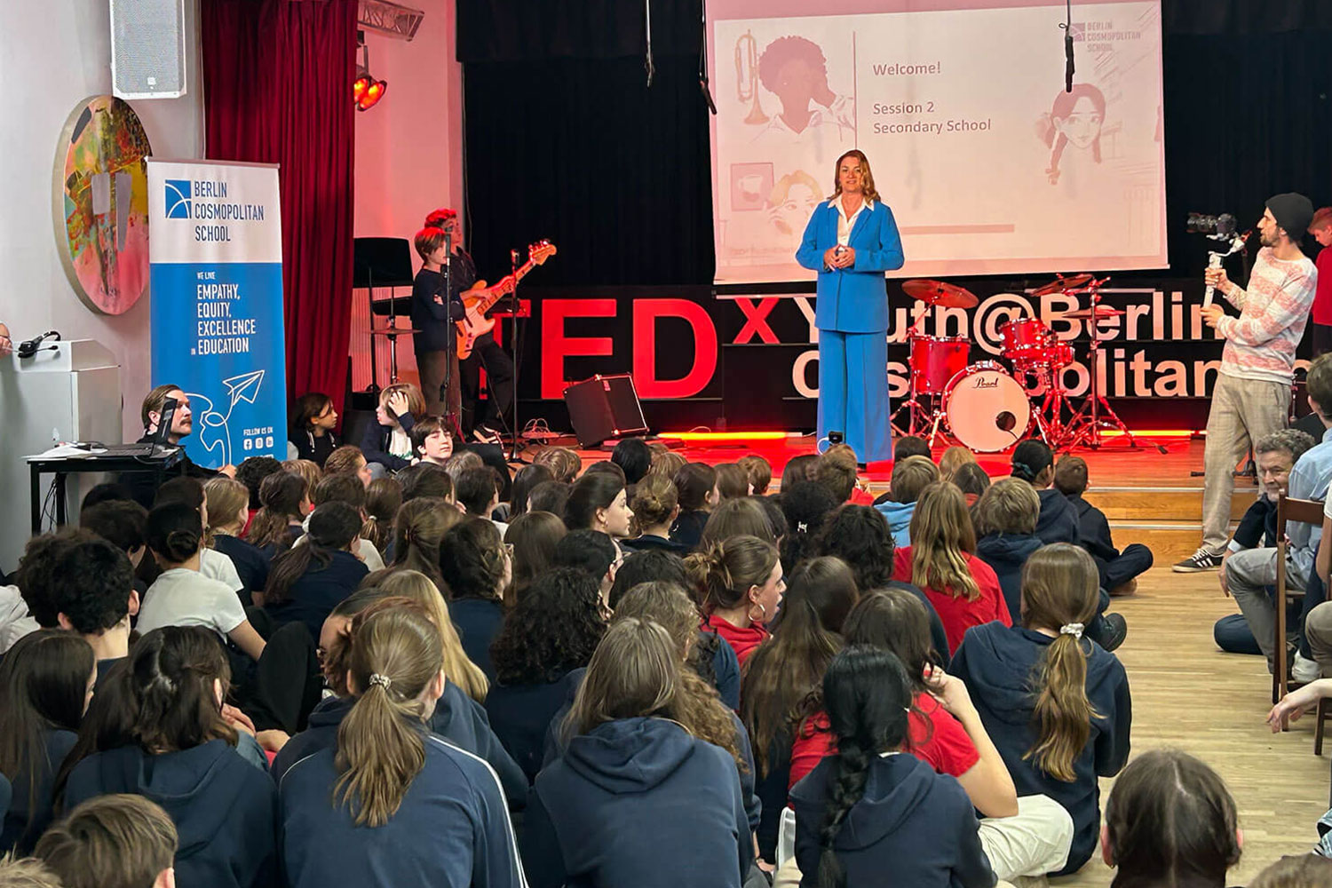 Cosmopolitanschool Berlin Eine Frau in einem blauen Anzug spricht auf der Bühne einer TEDx-Jugendveranstaltung in einer Schulaula. Auf dem Boden sitzen Schüler, Musiker, ein Kameramann und ein Banner der Berlin Cosmopolitan School ist zu sehen.