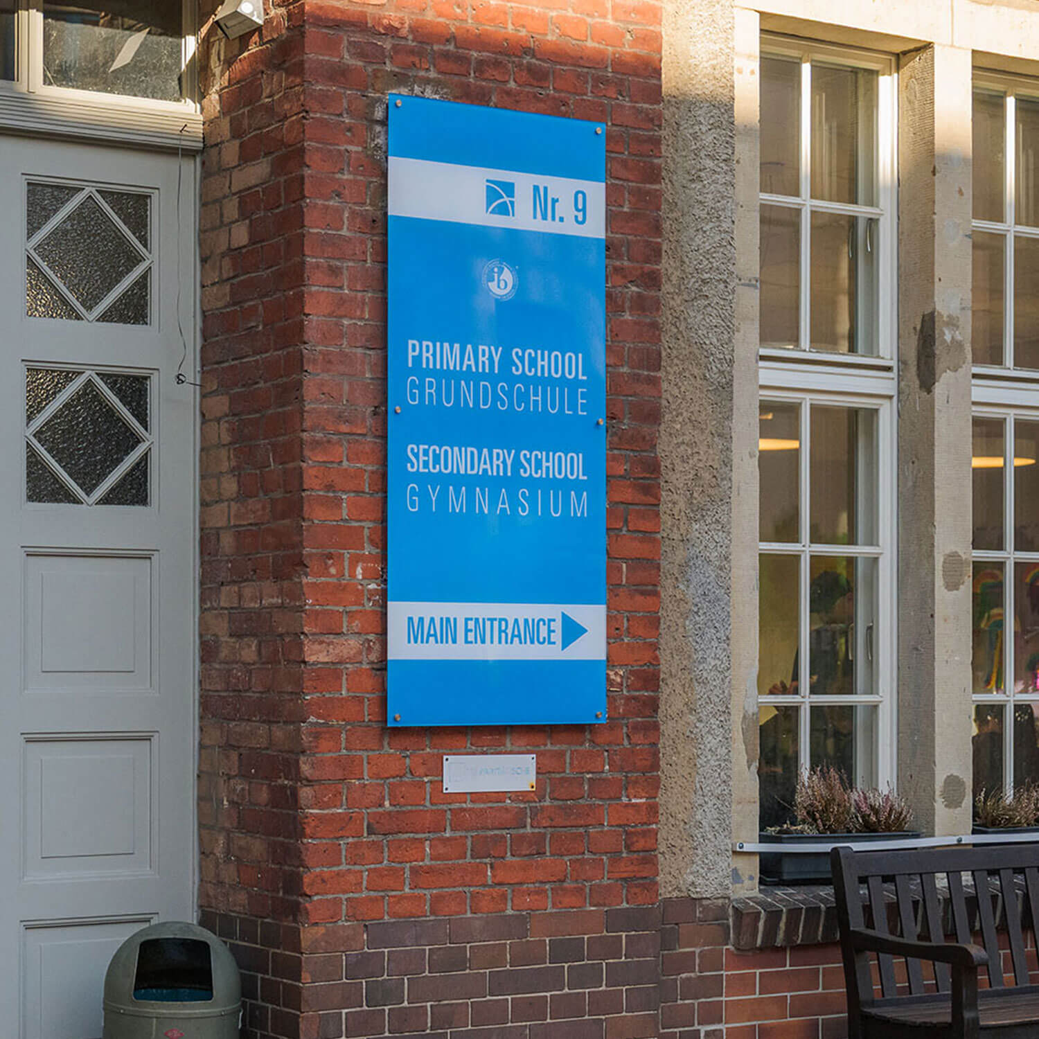 Cosmopolitanschool Berlin Auf einem blauen Schild an einem Backsteingebäude steht: Nr. 9, Kindergarten und Vorschule, Grundschule, Gymnasium, Haupteingang mit einem Pfeil, der nach rechts zeigt. Auf der linken Seite ist eine weiße Tür und auf der rechten Seite sind hohe Fenster. loading=