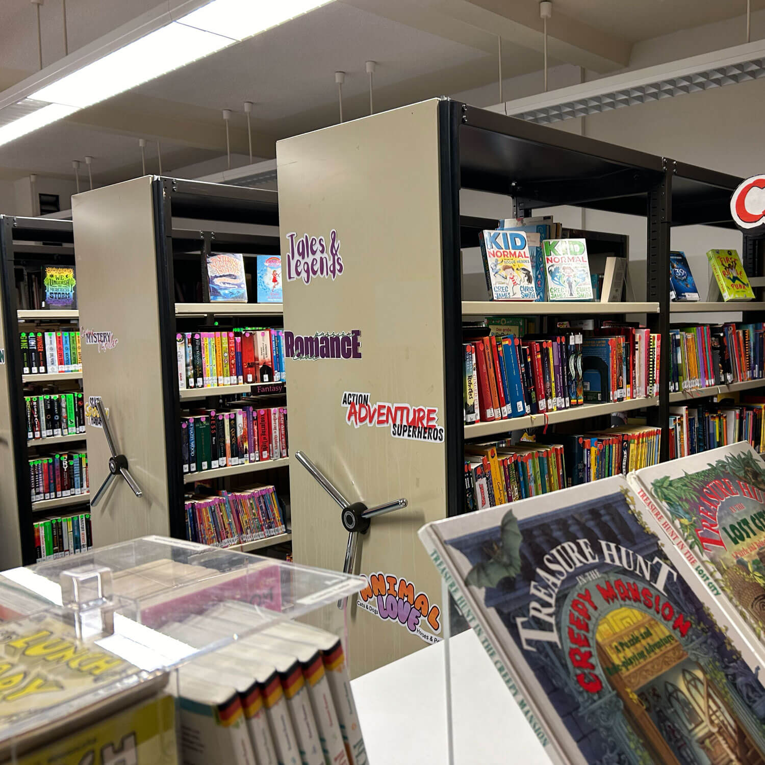 Cosmopolitanschool Berlin Eine Bibliothek mit Regalen mit farbenfrohen Kinderbüchern, beschriftet mit Rubriken wie "Märchen und Legenden", "Romantik", "Action-Abenteuer-Superhelden" und "Tierwelt". Im Vorder- und Hintergrund sind Buchumschläge zu sehen.