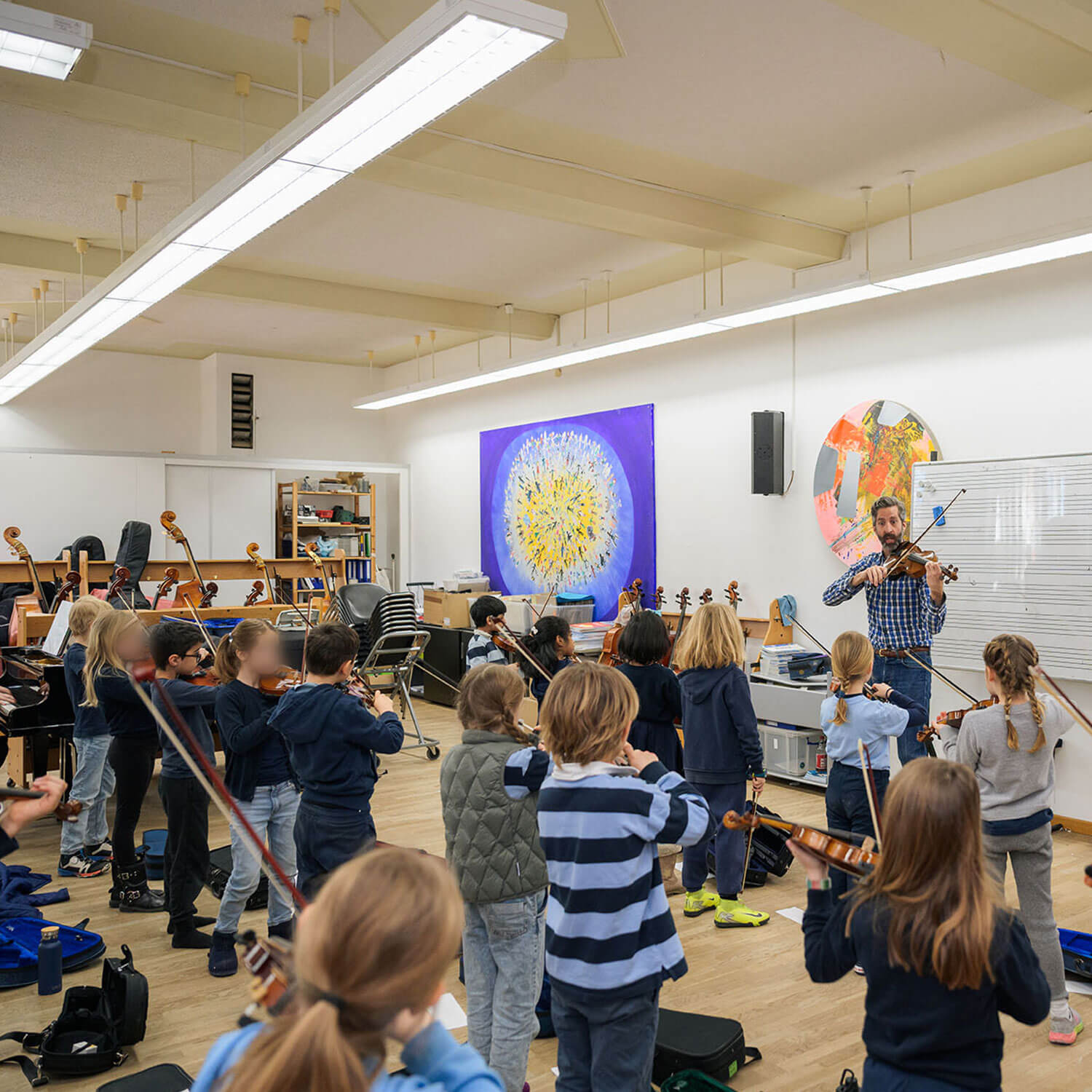 Cosmopolitanschool Berlin Eine Gruppe von Kindern spielt in einem Klassenzimmer Geige, während sie einem erwachsenen Lehrer gegenübersitzen. Der Raum ist hell beleuchtet, hat Notenständer und bunte Kunstwerke an den Wänden.