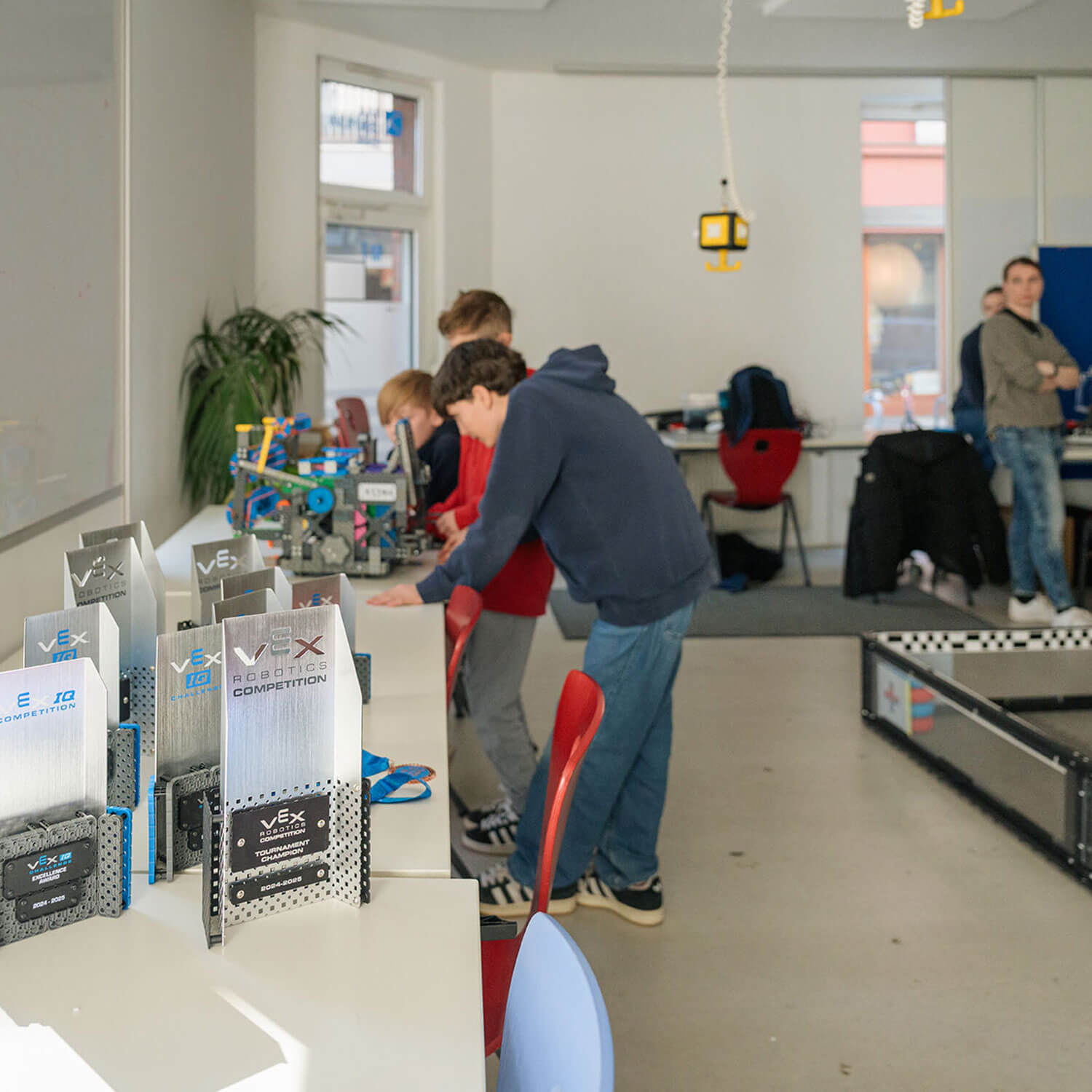 Cosmopolitanschool Berlin Eine Gruppe von Schülerinnen und Schülern arbeitet an einem Tisch in einem hellen Klassenzimmer an Robotikprojekten. Im Vordergrund sind die Trophäen des VEX-Wettbewerbs zu sehen, rechts eine Roboterarena.