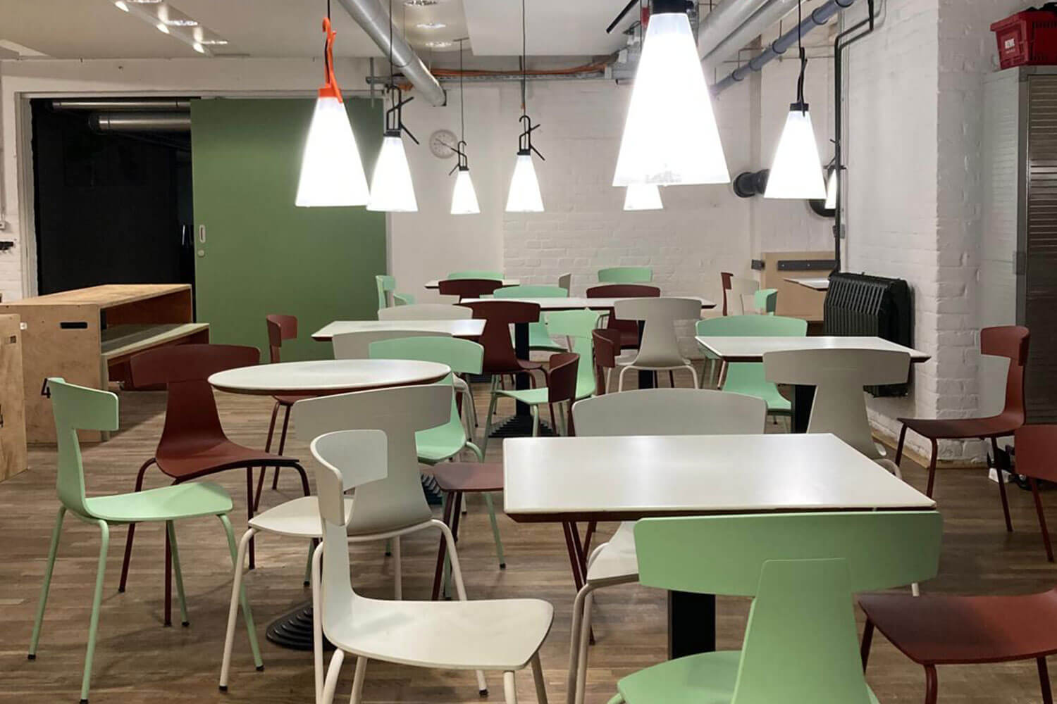 Cosmopolitanschool Berlin Ein modernes Café mit weißen Wänden, Holzböden und verschiedenen quadratischen und runden Tischen, umgeben von mintgrünen und roten Stühlen. Mehrere große Pendelleuchten hängen von der Decke und erhellen den leeren Raum.