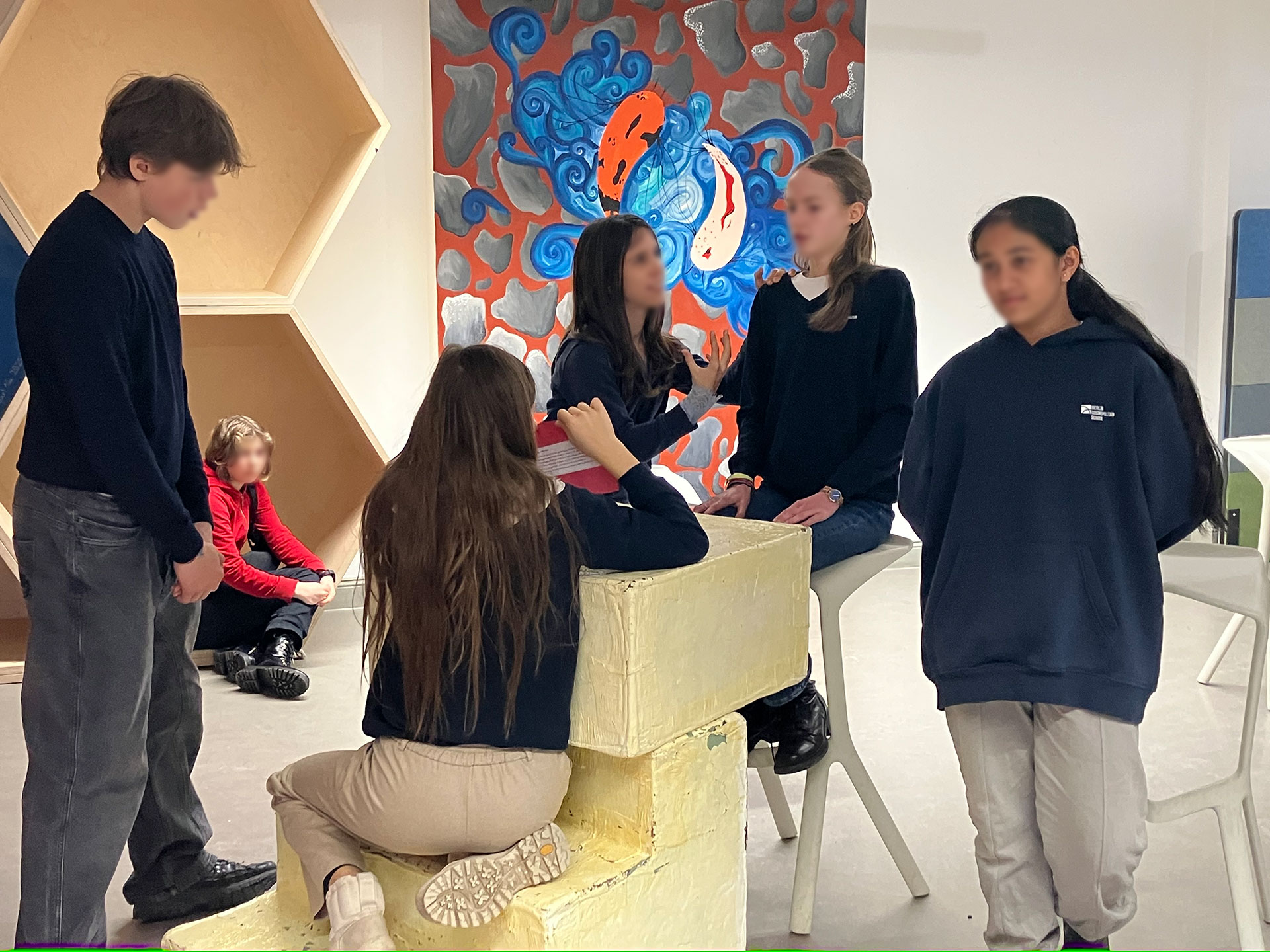 Cosmopolitanschool Berlin Eine Gruppe von Schülern der 9. Klasse in dunklen Pullovern und hellen Hosen unterhält sich und sitzt auf hellen Blöcken in einem Innenraum; ein Kind sitzt auf dem Boden im Hintergrund in der Nähe von Wandkunst mit blauen und orangen abstrakten Formen, während eines Englisch-Theaterworkshops.