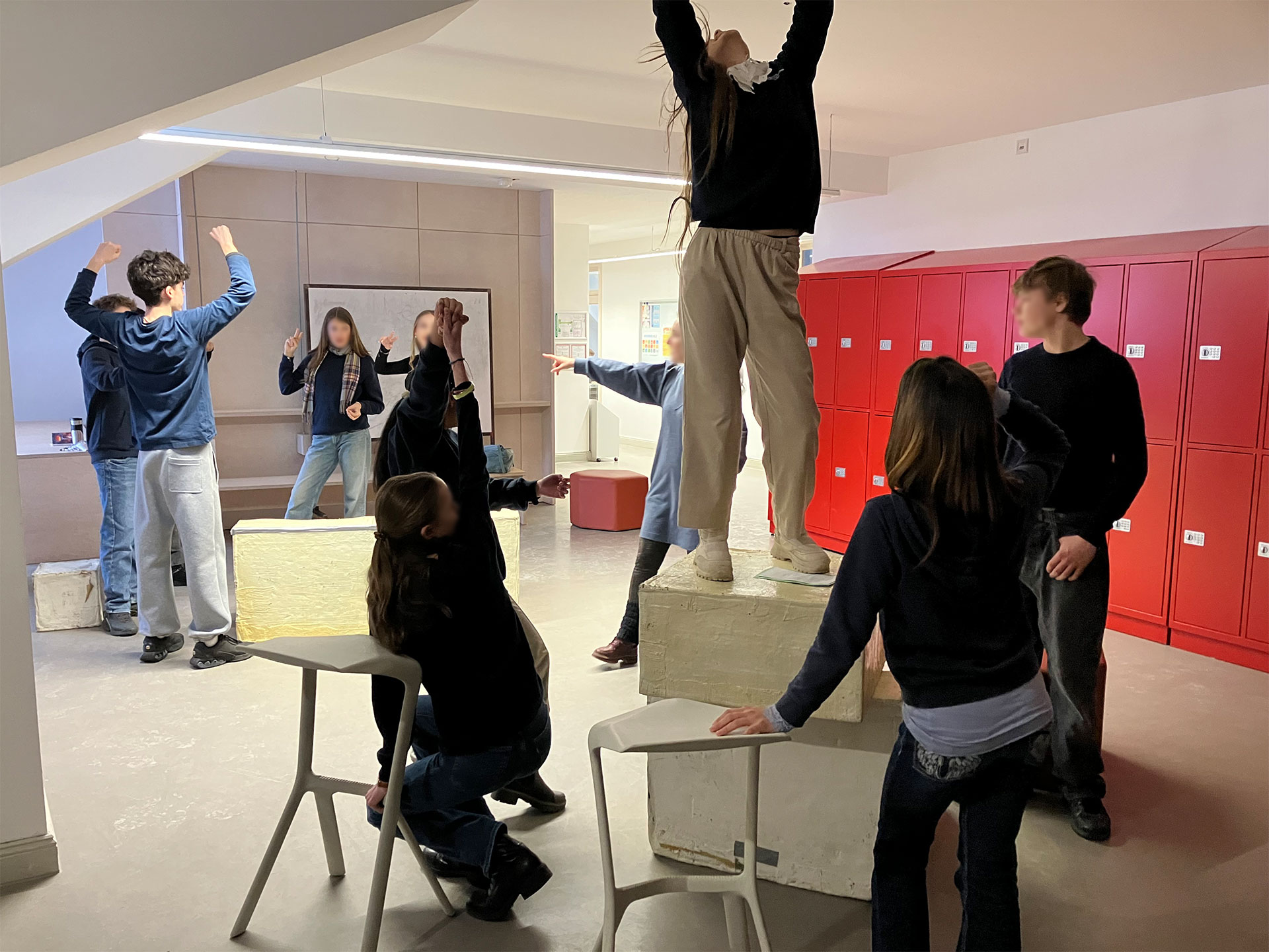 Cosmopolitanschool Berlin Eine Gruppe von Schülern der 9. Klasse in Freizeitkleidung posiert während eines Englisch-Theaterworkshops dynamisch in einem Raum mit roten Schließfächern, steht auf Bänken und Stühlen, einige heben die Arme, andere interagieren energisch.