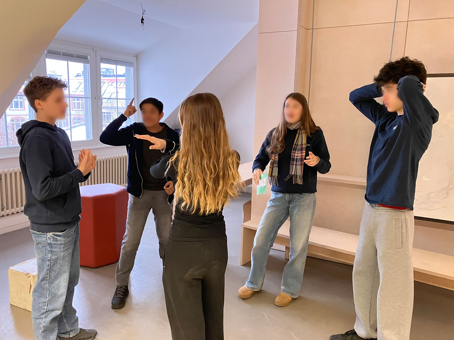 Cosmopolitanschool Berlin Fünf Jugendliche stehen in einem hellen Raum mit großen Fenstern, unterhalten sich angeregt und gestikulieren. Die Gruppe aus der 9. Klasse scheint in einen Englisch-Theaterworkshop oder eine Aktivität vertieft zu sein; eine Person steht mit dem Rücken zur Kamera.