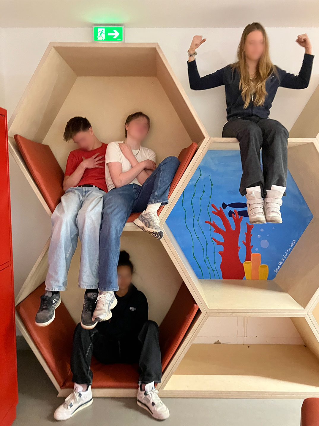 Cosmopolitanschool Berlin Vier Personen, wahrscheinlich aus einem Englisch-Theaterworkshop der 9. Klasse, sitzen und posieren in einem wabenförmigen Holzregal; zwei auf der linken Seite, eine darunter und eine darüber, die die Arme hebt. Ihre Gesichter sind unscharf; rechts ist eine Unterwasserszene gemalt. loading=
