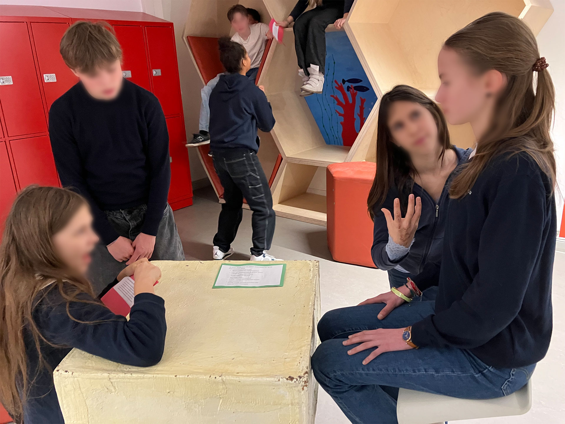 Cosmopolitanschool Berlin Eine Gruppe von Schülerinnen und Schülern der 9. Klasse in einem modernen Klassenzimmer. Einige unterhalten sich an einem Tisch, während andere während eines Englisch-Theaterworkshops an roten Spinden stehen und auf wabenförmige Holzregale klettern.
