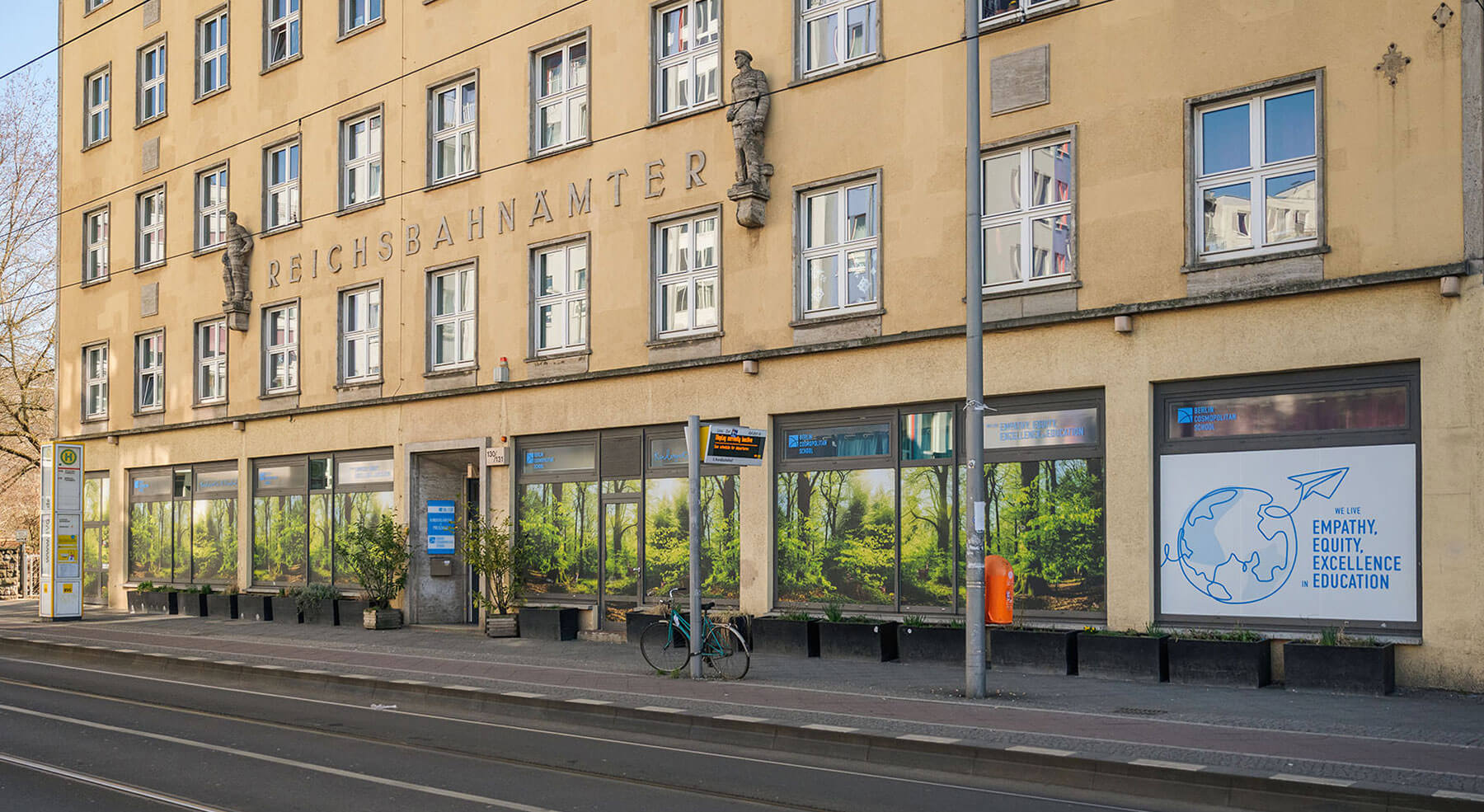 Cosmopolitanschool Berlin Ein beigefarbenes Gebäude mit der Aufschrift REICHSBAHNAMTER, mit großen Fenstern, einer Straßenbahnhaltestelle, Bänken, einem grünen Fahrrad, Bäumen, die sich im Glas spiegeln, und einem Plakat mit einer Weltkugel und den Worten Empathie, Gerechtigkeit, Exzellenz - ideal für Kindergarten und Vorschule.