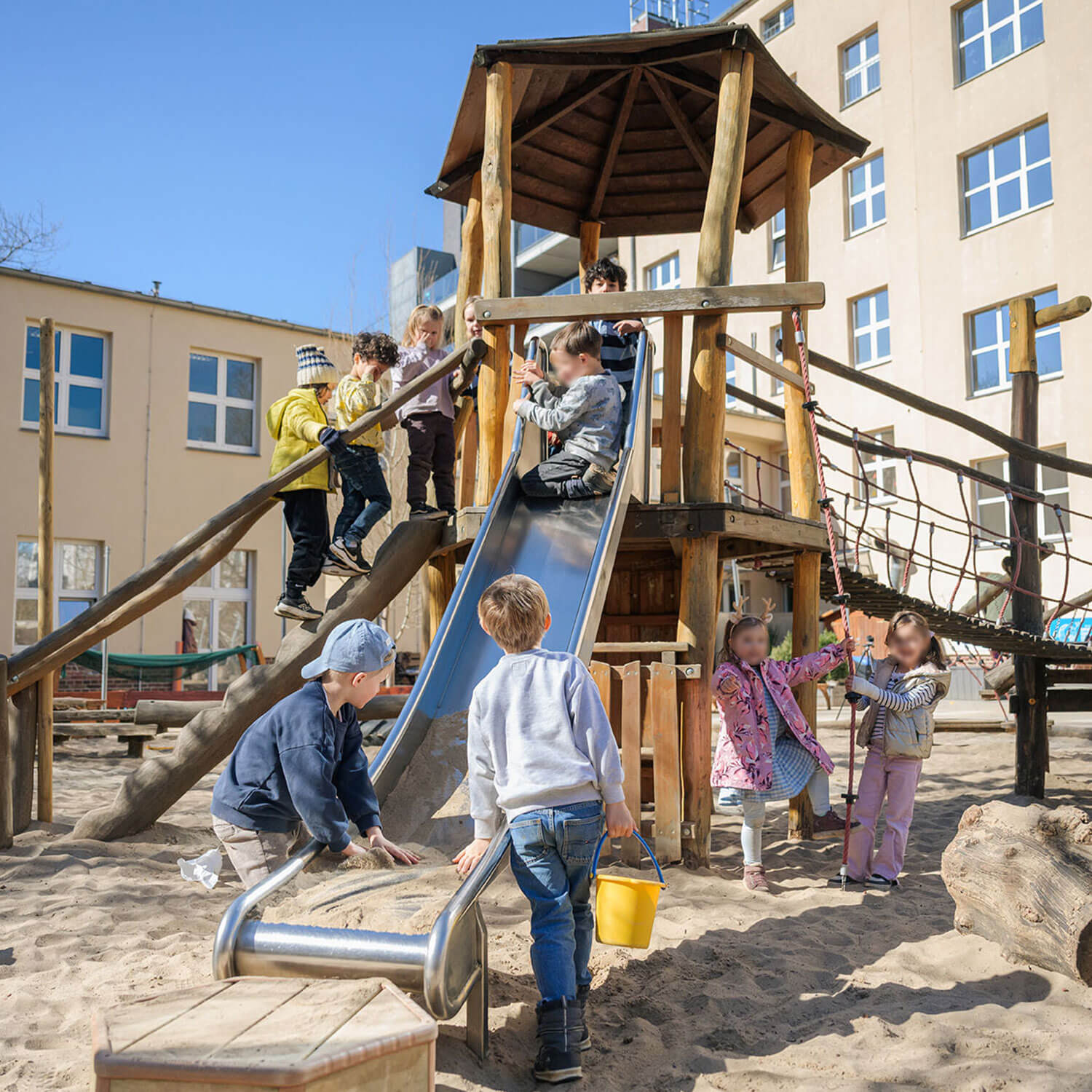 Cosmopolitanschool Berlin Eine Gruppe von Kindern spielt auf einem hölzernen Spielplatz mit einer Rutsche und Kletternetzen in einem Kindergarten und einer Vorschule, umgeben von Sand. Einige klettern die Stufen hinauf, andere warten unten, und wieder andere schauen zu oder spielen in der Nähe. Im Hintergrund sind Gebäude zu sehen.