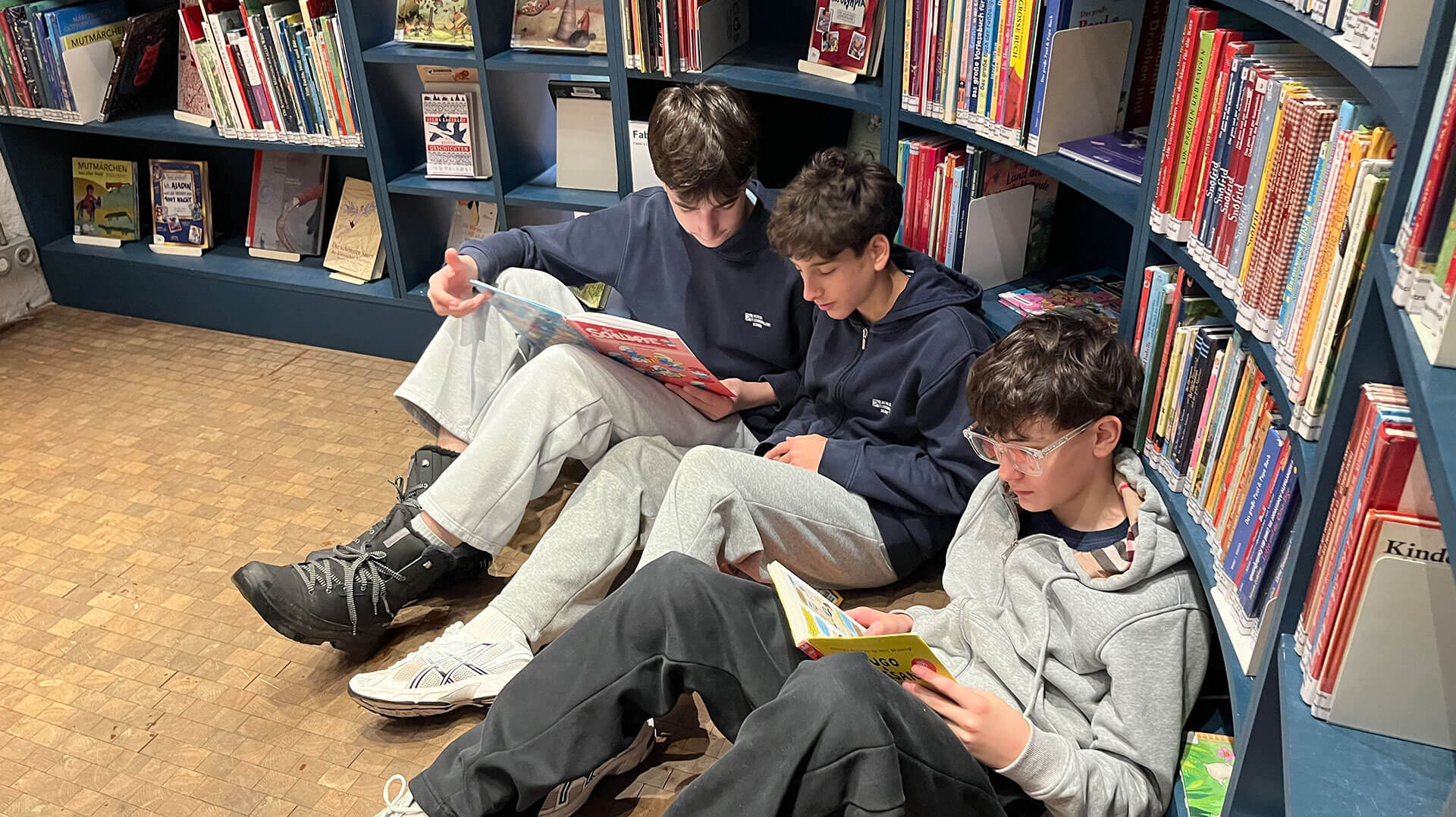 Cosmopolitanschool Berlin Drei Jungen aus den 7. Klassen sitzen auf dem Boden der Philip-Schäfer-Bibliothek und lesen Bücher. Umgeben von bunten Regalen wirken sie konzentriert und entspannt in dieser gemütlichen Lesereise durch Geschichten und Fantasie. loading=