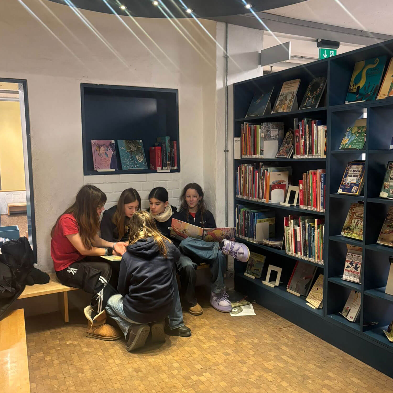 Cosmopolitanschool Berlin Fünf Mädchen aus der 7. Klasse sitzen in einer gemütlichen Ecke der Philip-Schäfer-Bibliothek zusammen und lesen und erkunden Bücher in dunkelblauen Regalen - der perfekte Ort für ihre Lesereise. loading=