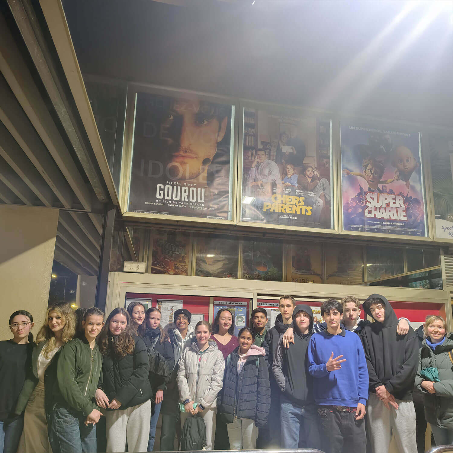 Cosmopolitanschool Berlin Eine Gruppe von Neuntklässlern posiert gemeinsam vor einem nächtlichen Kino. Sie stehen unter hell erleuchteten Plakaten für drei Filme, lächeln und machen lässige Gesten. loading=