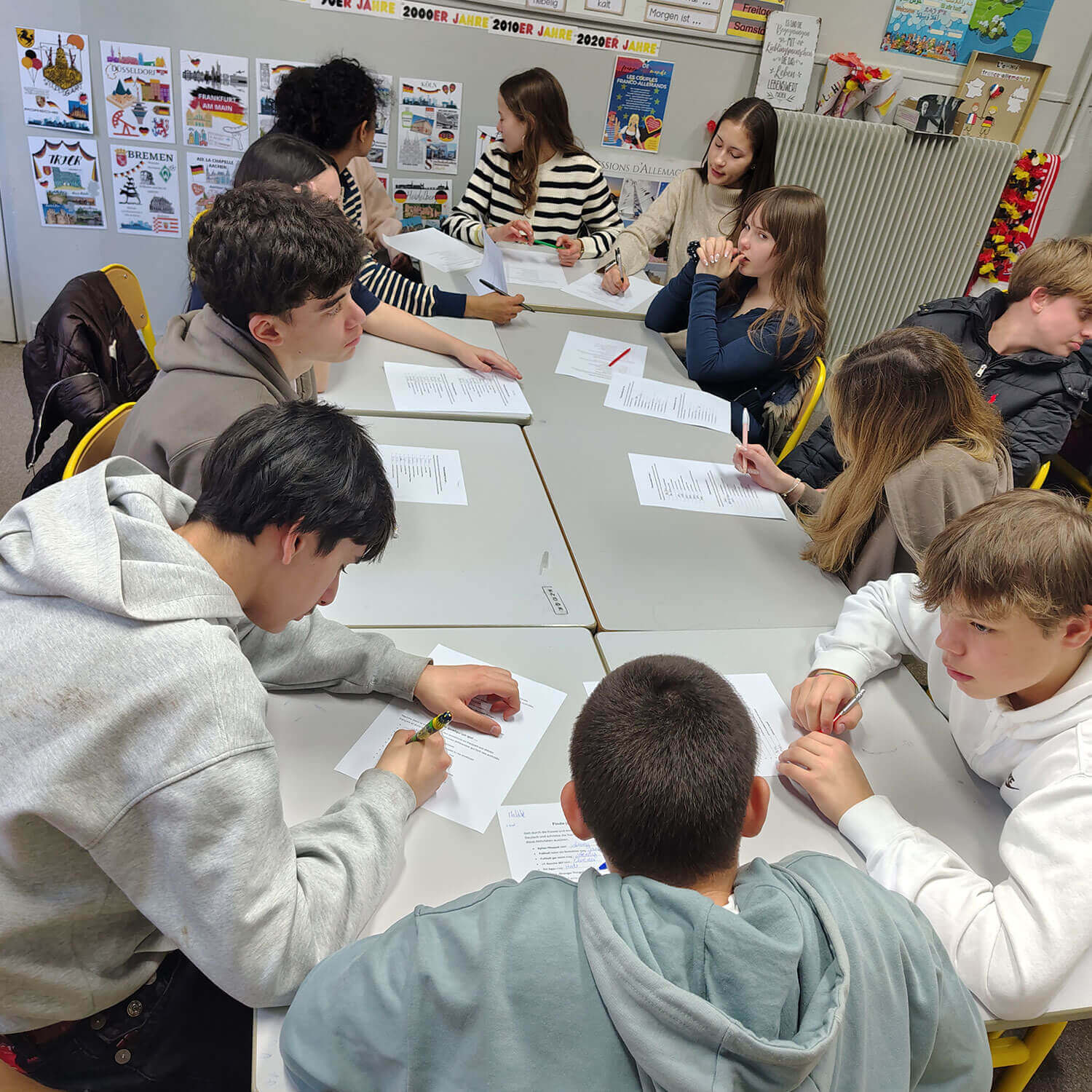 Cosmopolitanschool Berlin Eine Gruppe von Neuntklässlern sitzt in einem Klassenzimmer an Tischen, arbeitet gemeinsam an Aufgaben und bespricht Referate. Die Wände sind mit bunten Postern, einem Kalender und sogar Hinweisen auf berühmte Märchen geschmückt.