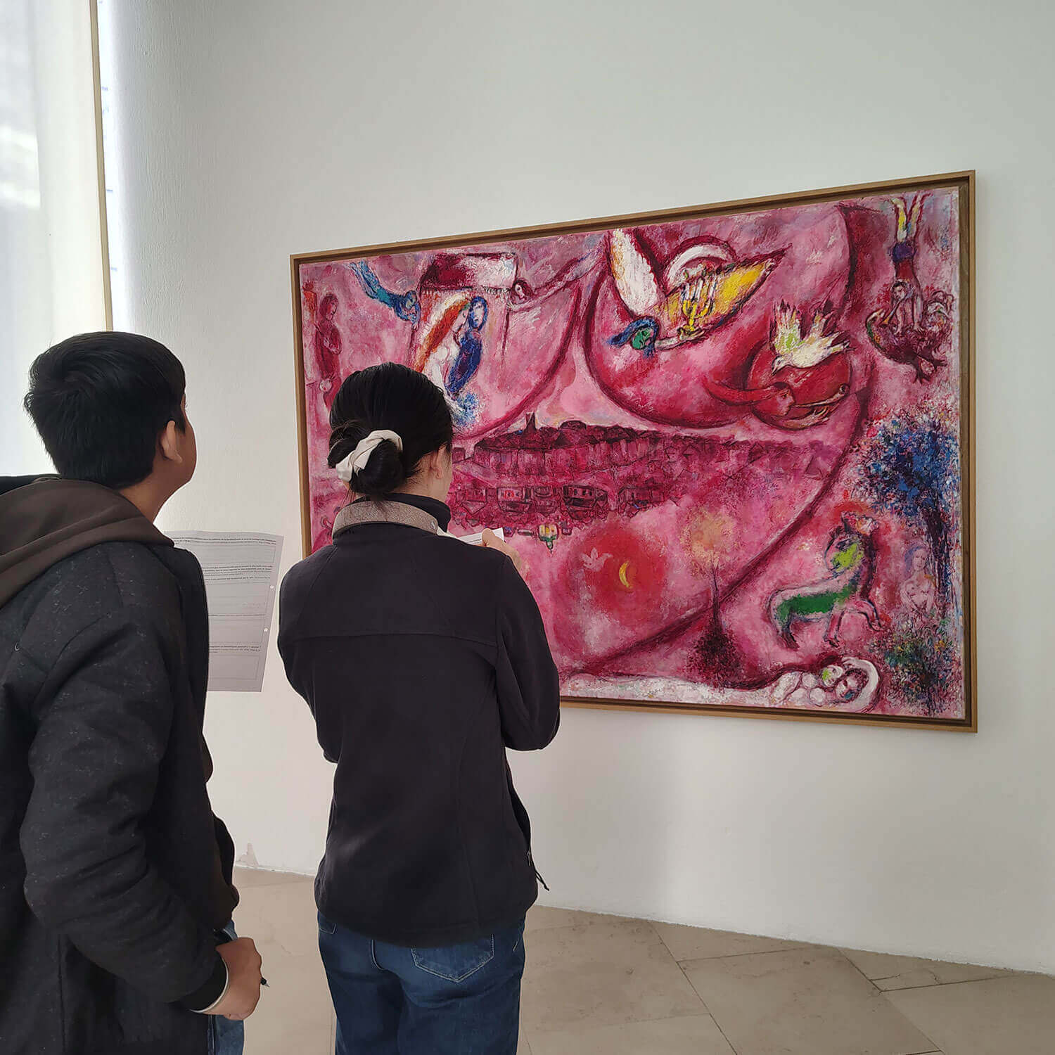 Cosmopolitanschool Berlin Zwei Personen stehen vor einem leuchtenden, rosafarbenen Gemälde mit abstrakten und phantastischen Elementen, die an ein Märchen erinnern, und betrachten und diskutieren dessen Details in einer Galerie.