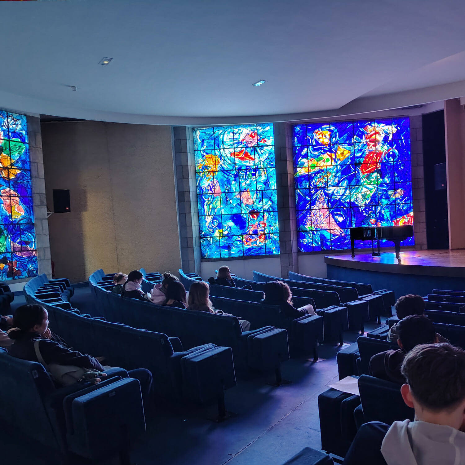 Cosmopolitanschool Berlin Die Zuschauer sitzen in einem schwach beleuchteten Auditorium mit blauen Sitzreihen und blicken auf eine Bühne, auf der ein Flügel unter leuchtenden Glasfenstern in Côte d'Azur-Farben steht, die mit bunten Mustern akzentuiert sind, die an ein Märchen erinnern.