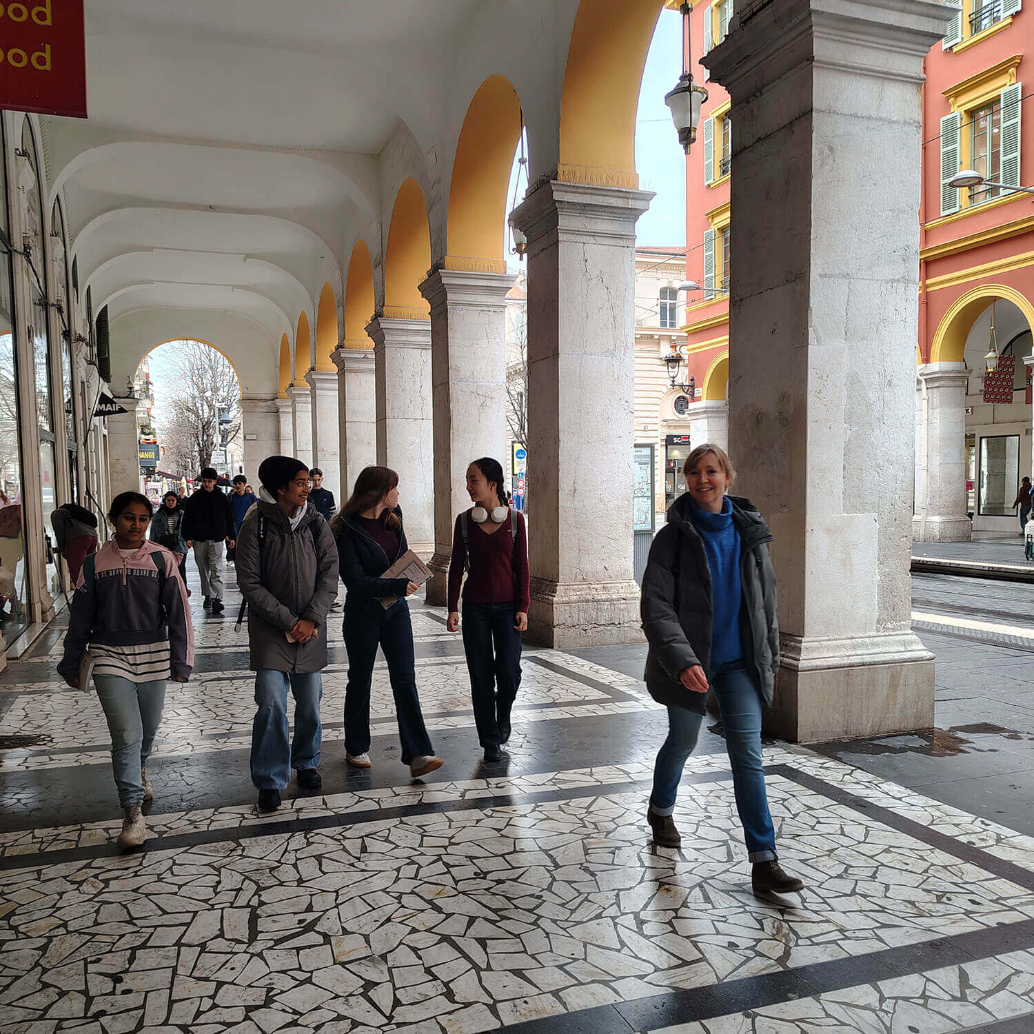 Cosmopolitanschool Berlin Menschen gehen unter einem gewölbten, überdachten Gang mit hohen Säulen und einem gefliesten Boden in einer Stadt, die an die Côte d'Azur erinnert. Im Hintergrund sind bunte Gebäude und eine belebte Straße zu sehen, die eine lockere Atmosphäre schaffen.