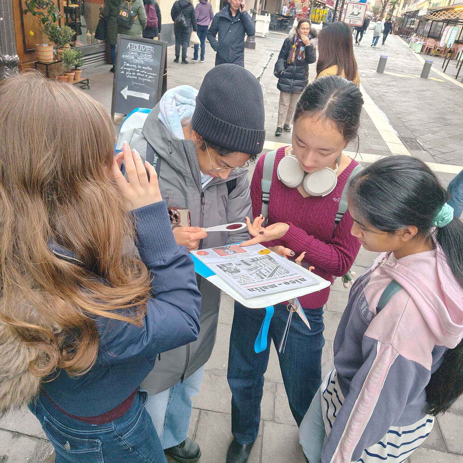 Cosmopolitanschool Berlin Vier junge Menschen, möglicherweise Neuntklässler, stehen dicht beieinander auf einer Straße in der Stadt und betrachten eine gedruckte Karte. Einer hält die Karte in der Hand, während die anderen aufmerksam auf sie zeigen und ihre nächsten Schritte planen - vielleicht träumen sie von einer Reise an die Côte d'Azur.