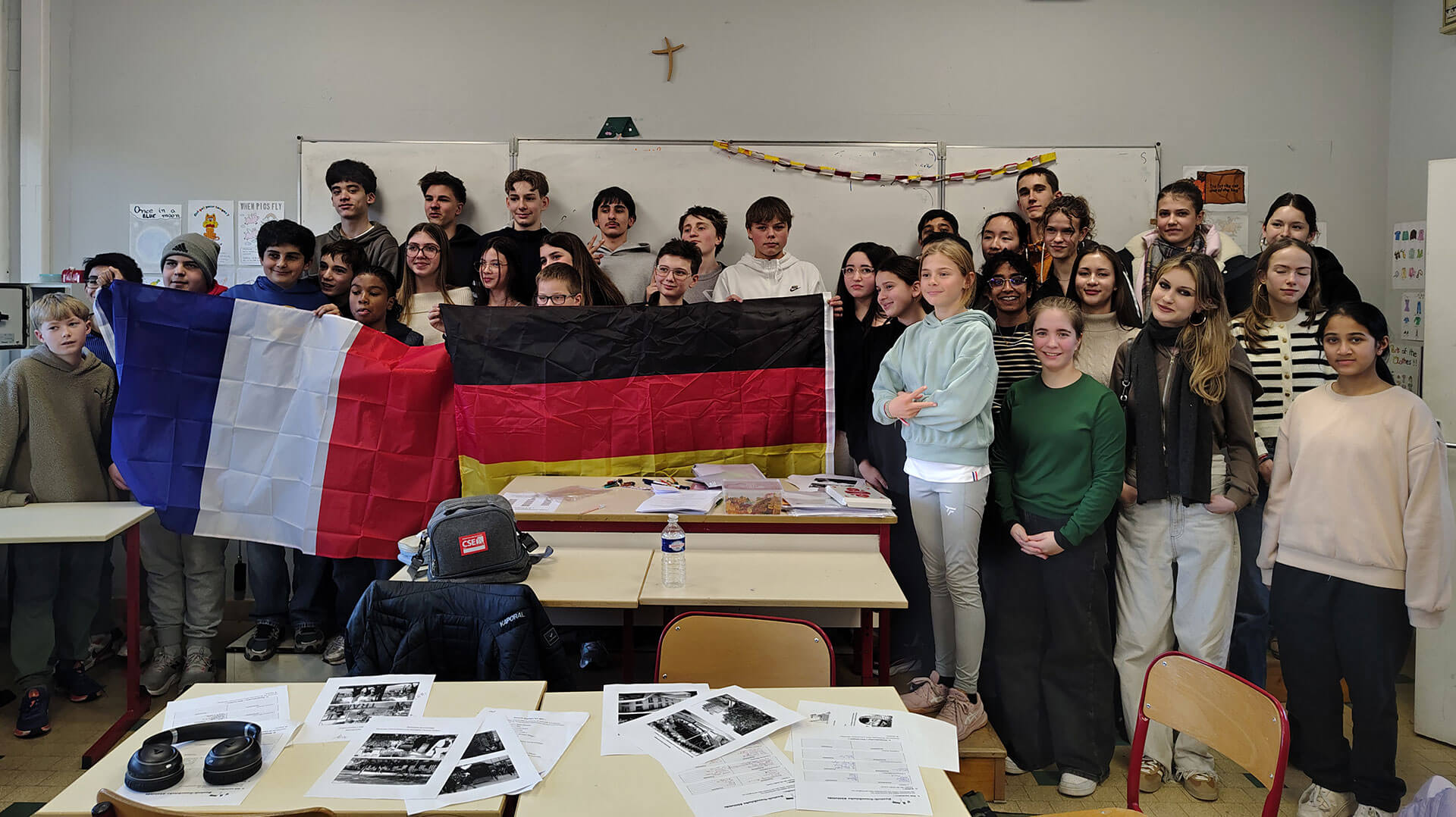 Cosmopolitanschool Berlin Eine Gruppe von Schülern posiert in einem Klassenzimmer und hält große französische und deutsche Fahnen in die Höhe. Auf den Tischen davor liegen Papiere, und an der Wand dahinter sind ein Kreuz und Dekorationen zu sehen. loading=