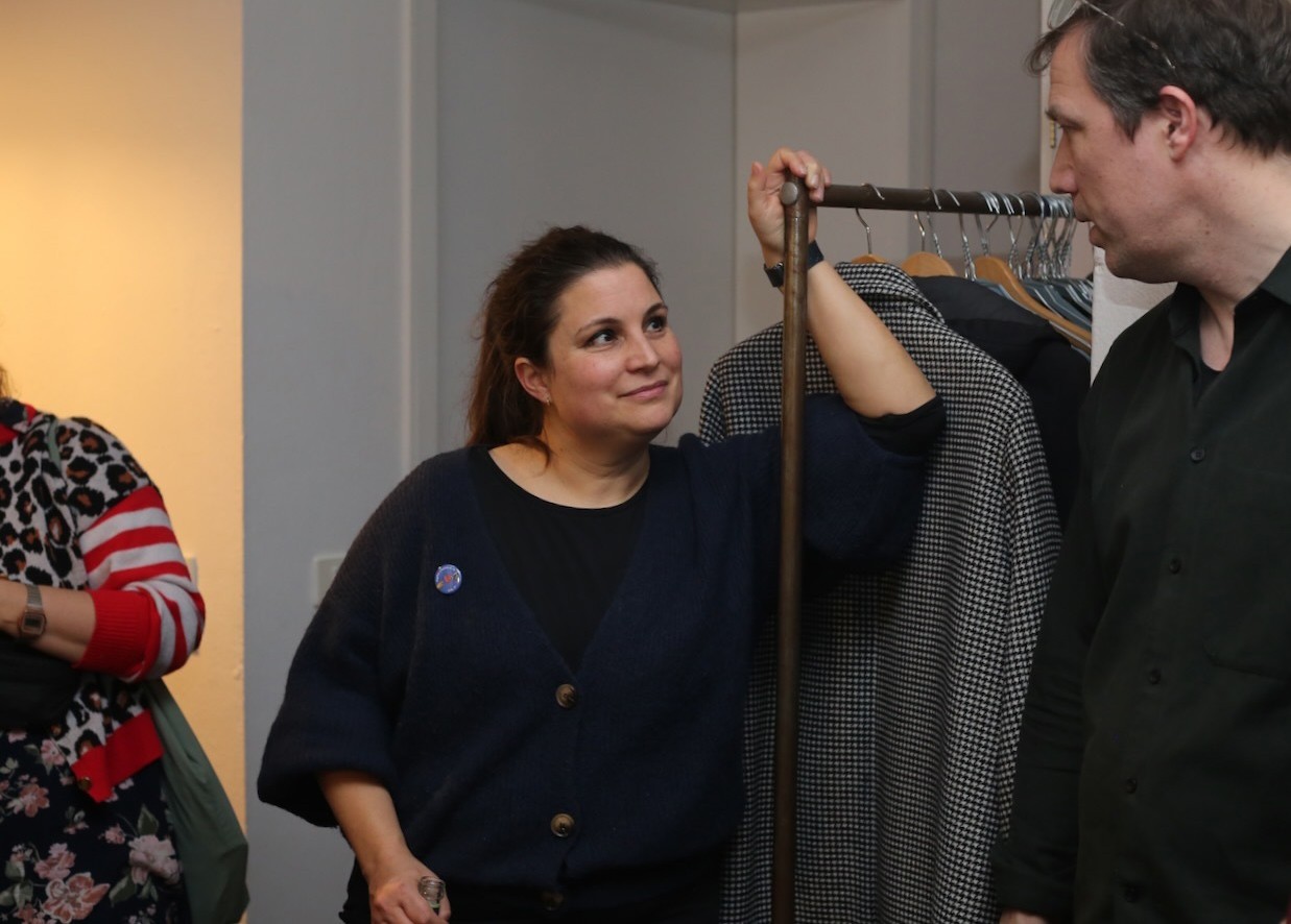 Cosmopolitanschool Berlin Eine Frau lehnt an einem Kleiderständer, lächelt und blickt einen Mann in einem dunklen Hemd an. Eine weitere Person in gemusterter Kleidung steht in der Nähe, teilweise außerhalb des Bildausschnitts. Die warme Innenraumatmosphäre suggeriert einen nachdenklichen Moment über Verantwortung übernehmen. loading=