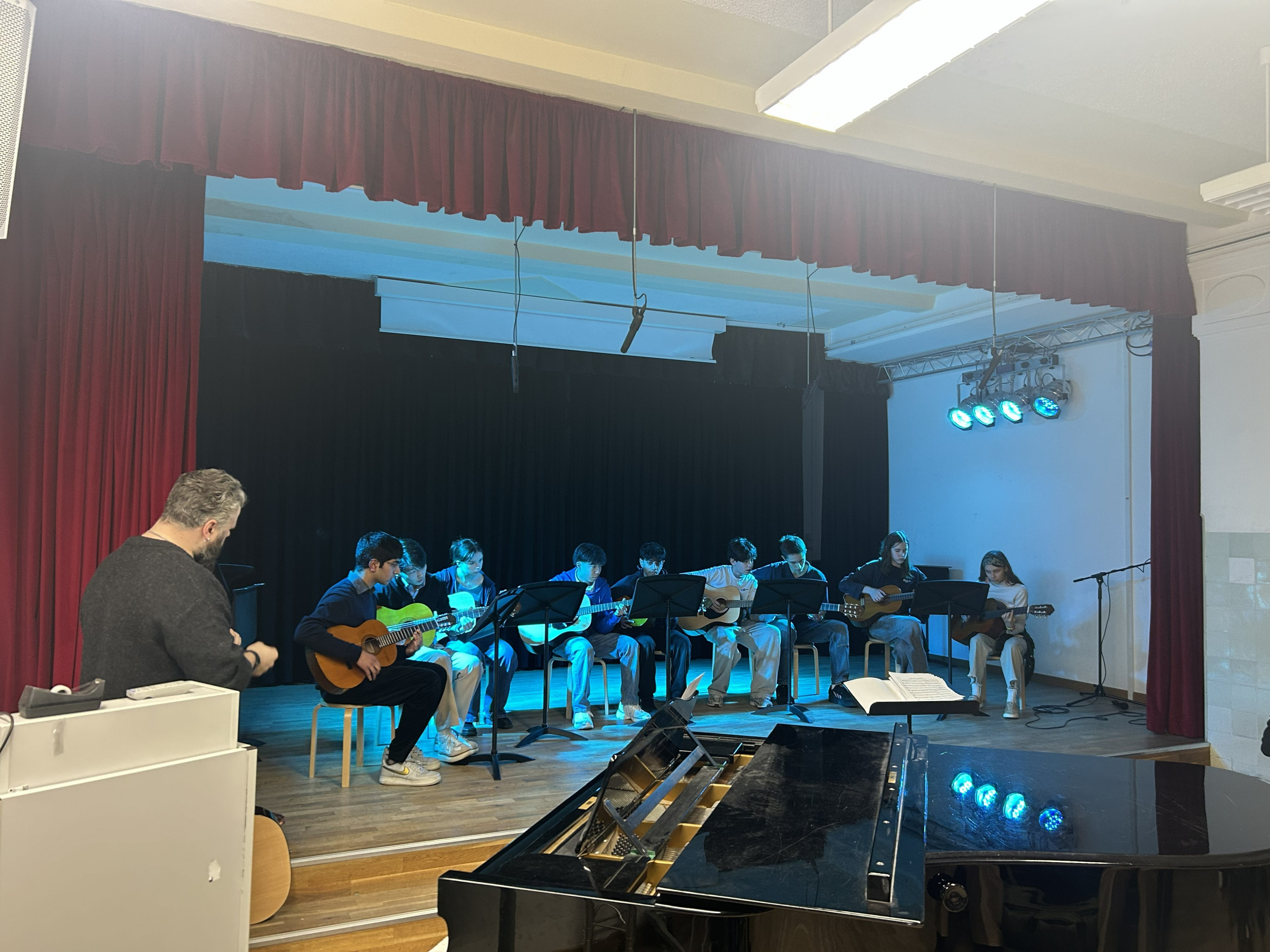 Cosmopolitanschool Berlin Eine Gruppe von Kindern sitzt auf der Bühne und spielt Gitarre, während ein Erwachsener an der Seite steht und zusieht. Die Bühne ist mit blauen Lichtern beleuchtet, und im Vordergrund ist ein Klavier zu sehen. Rote Vorhänge rahmen die Szene ein.