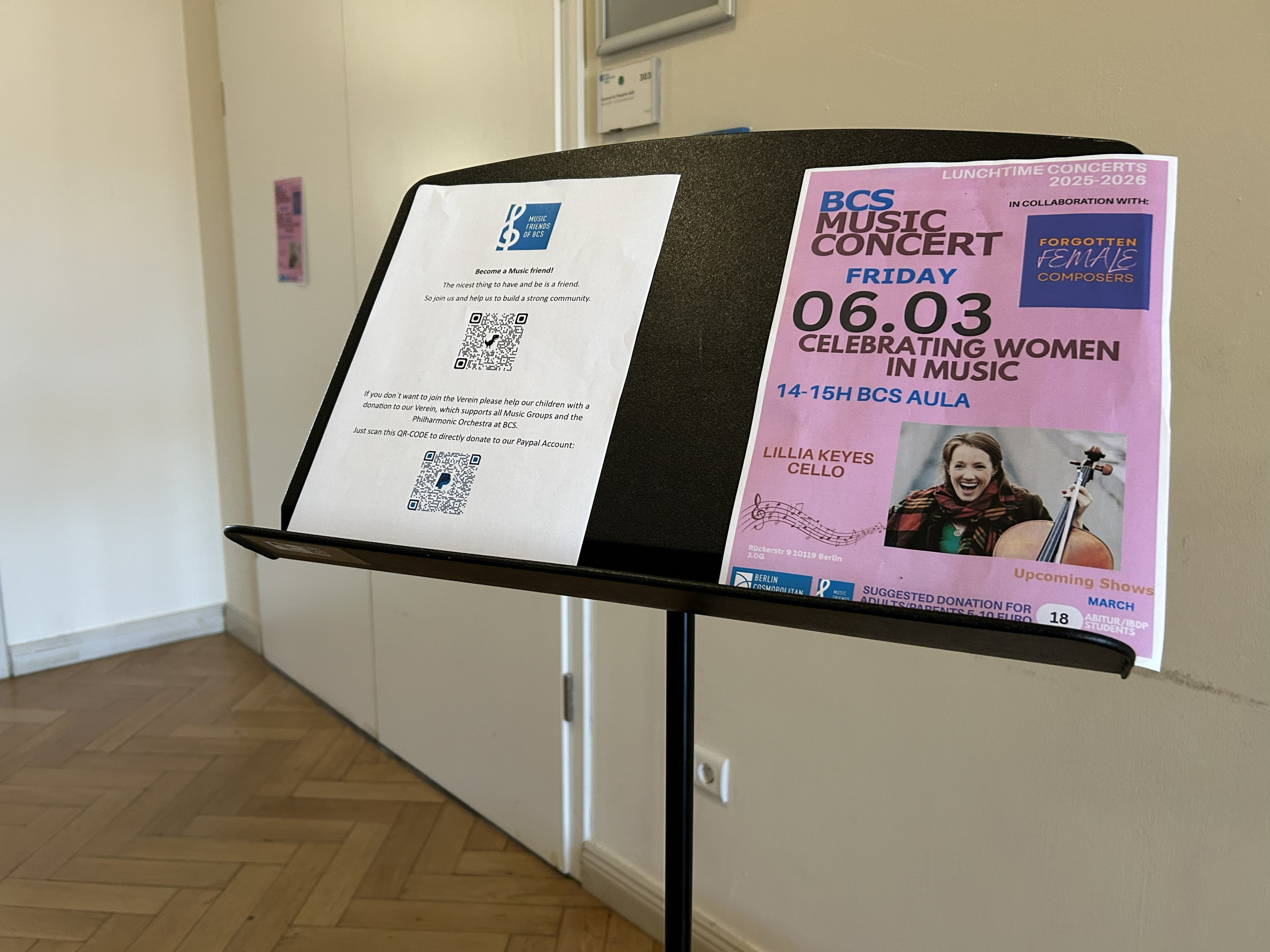 Cosmopolitanschool Berlin Ein schwarzer Notenständer hält zwei Papiere: eine gedruckte Notiz mit QR-Codes und ein buntes Plakat für ein BCS-Musikkonzert am 06.03. mit der Cellistin Lilla Keyes, das Frauen in der Musik feiert.