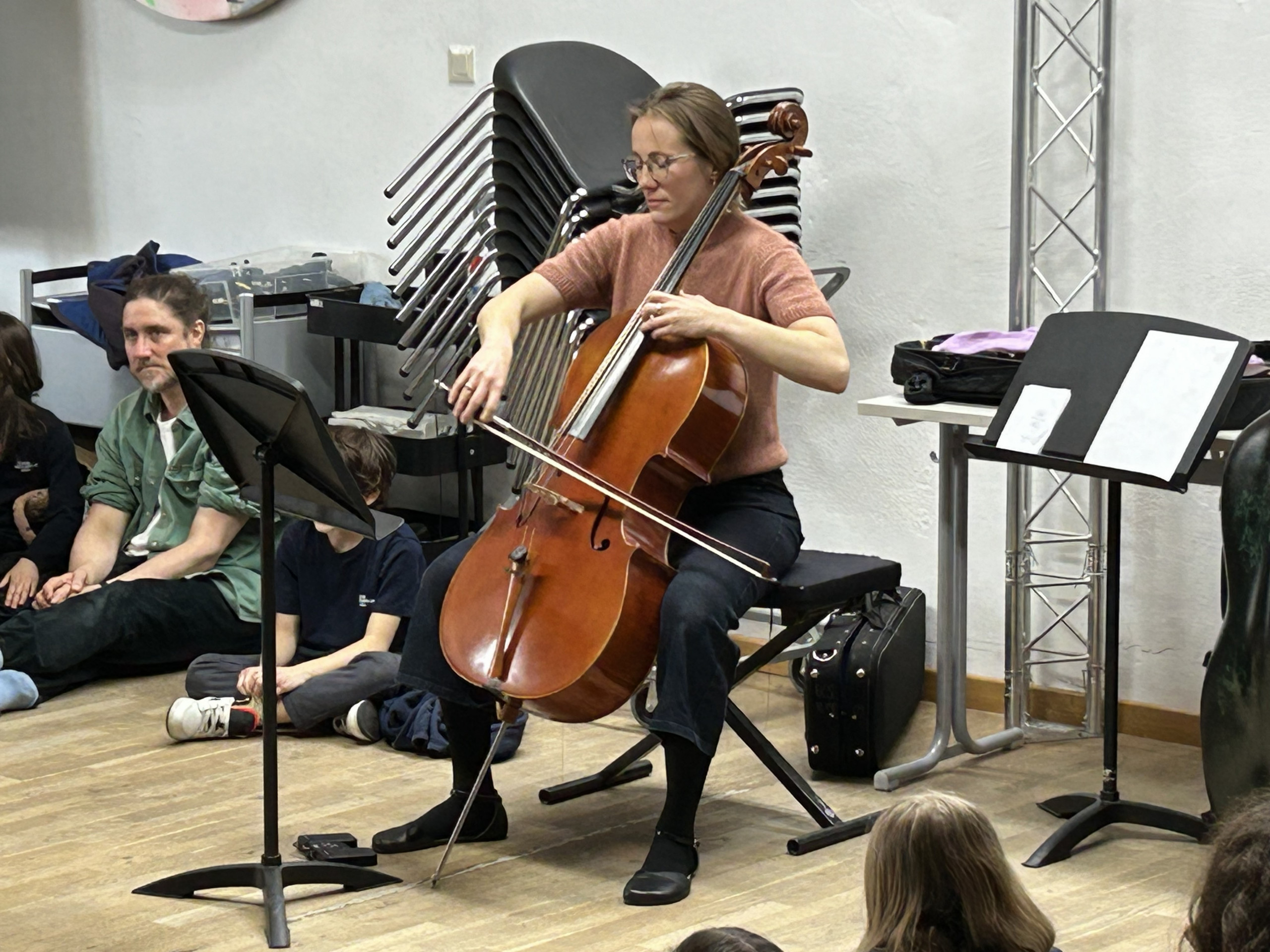 Cosmopolitanschool Berlin Eine Frau spielt Cello, während sie in einem Raum sitzt und ein Publikum von Kindern und Erwachsenen um sie herum auf dem Boden sitzt. Im Hintergrund sind gestapelte Stühle und Notenblätter zu sehen.