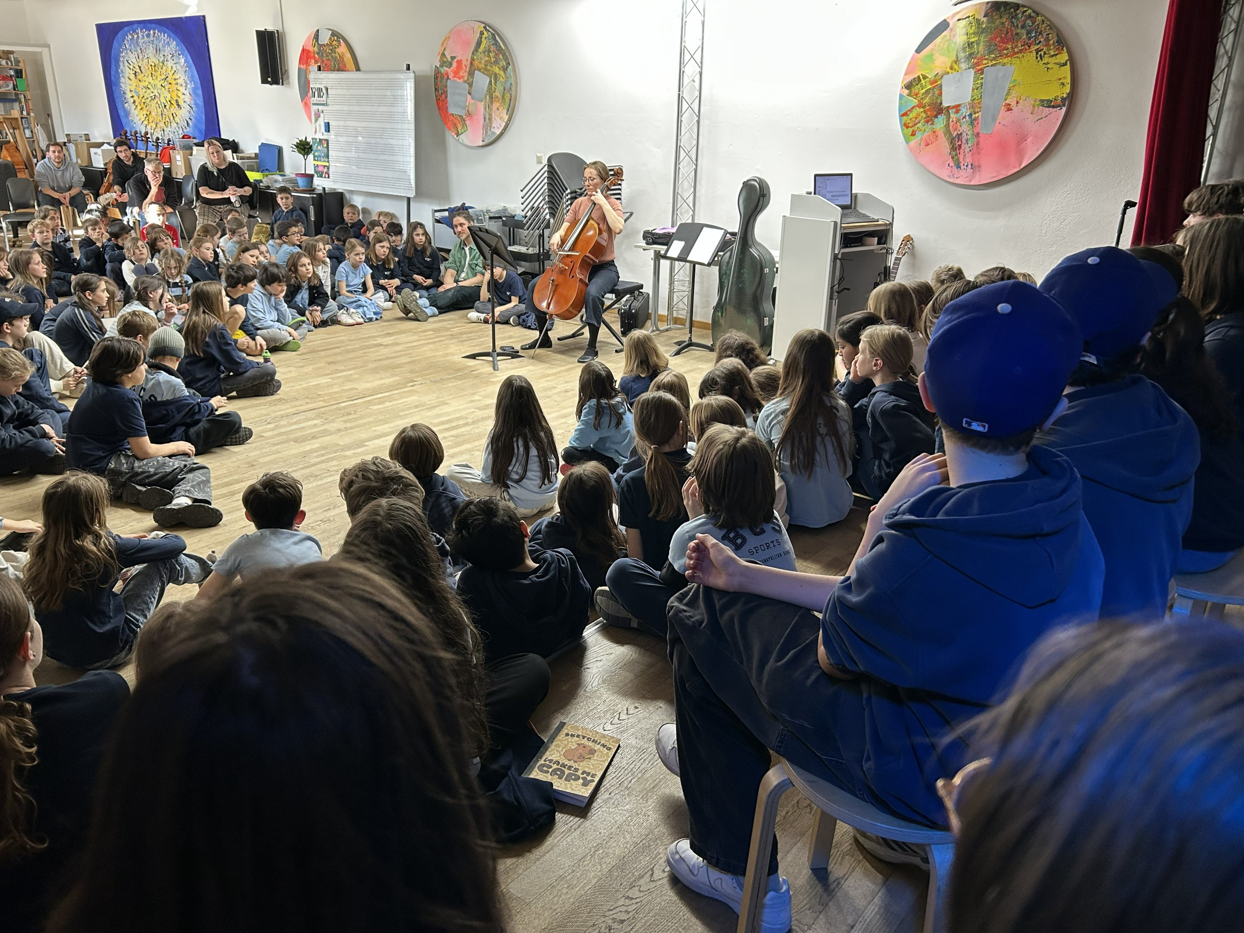 Cosmopolitanschool Berlin Ein Cellist spielt vor einem großen Publikum von Kindern, die auf dem Boden und auf Stühlen in einem hellen Raum mit bunten Kunstwerken an den Wänden sitzen. Das Publikum schaut aufmerksam zu.