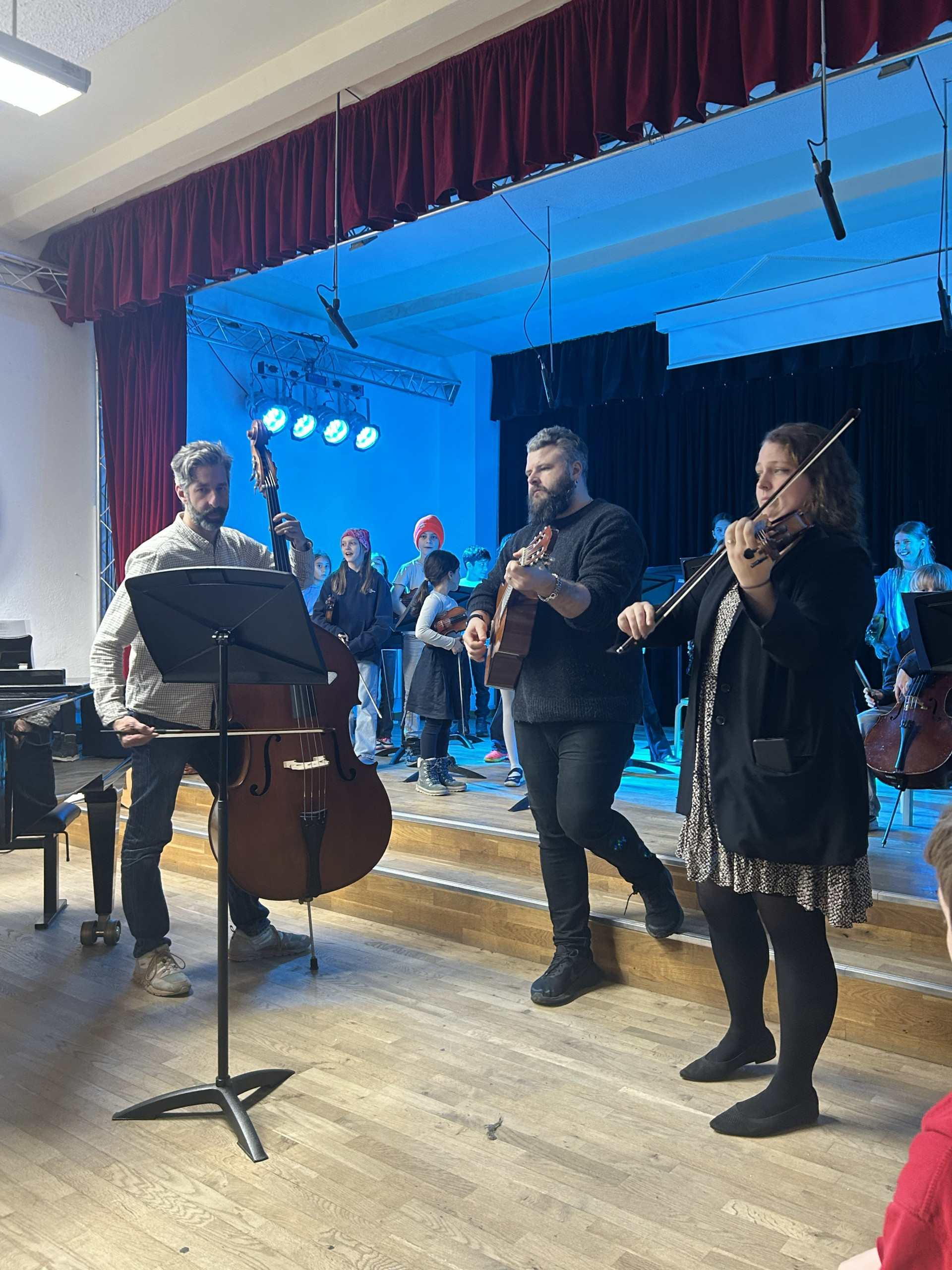 Cosmopolitanschool Berlin Drei Musiker treten auf einer Holzbühne auf: ein Kontrabassist, ein Akustikgitarrist und ein Geiger. Hinter ihnen singt ein Kinderchor unter blauem Bühnenlicht und einem roten Vorhang. loading=