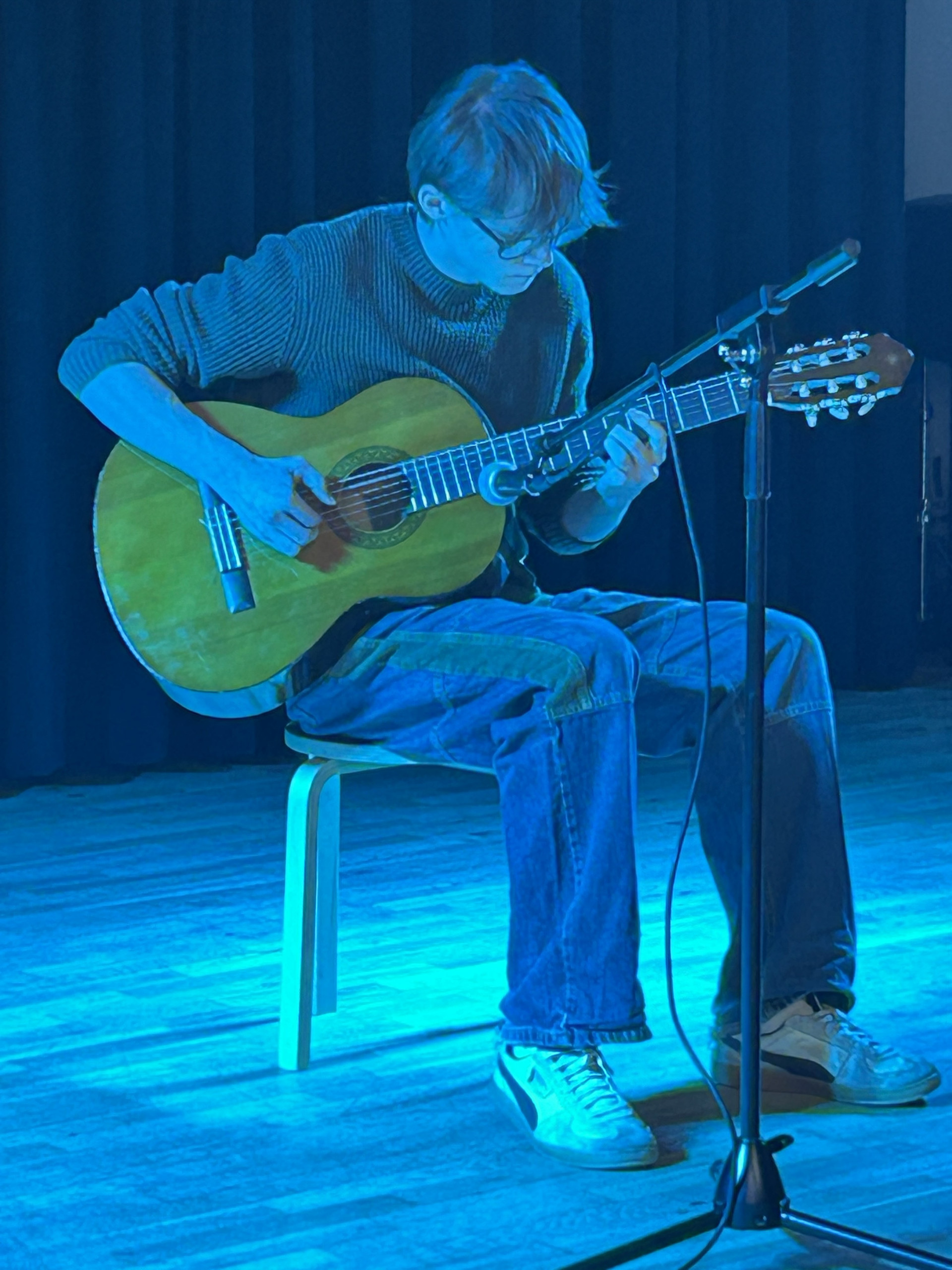 Cosmopolitanschool Berlin Eine Person mit hellem Haar und Brille sitzt auf einem Hocker und spielt auf der Bühne unter blauem Licht eine Akustikgitarre, vor ihr steht ein Mikrofonständer. loading=